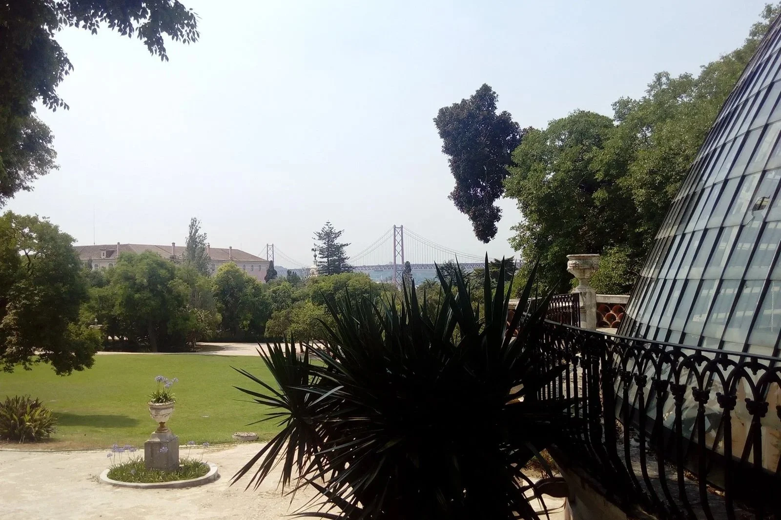 View of a lush green garden with trees, plants, and a decorative urn in the foreground, a cityscape with residential buildings, a bridge in the distance, and a modern glass building on the right side under a cloudy sky.