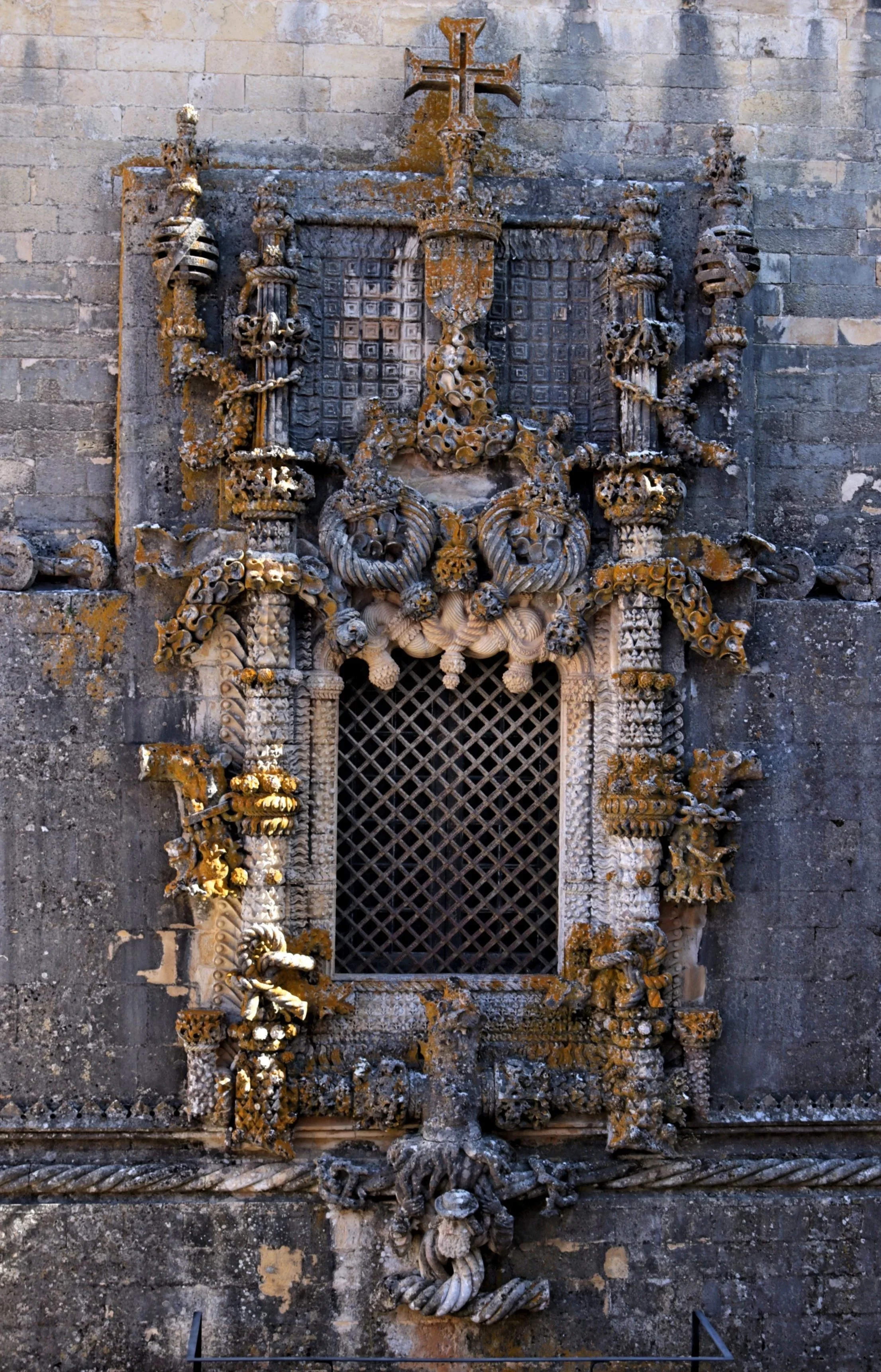 An ornate stone religious altar or monument against a stone wall, featuring a cross at the top, detailed carvings of flora and fauna, and a small grated window at the bottom.