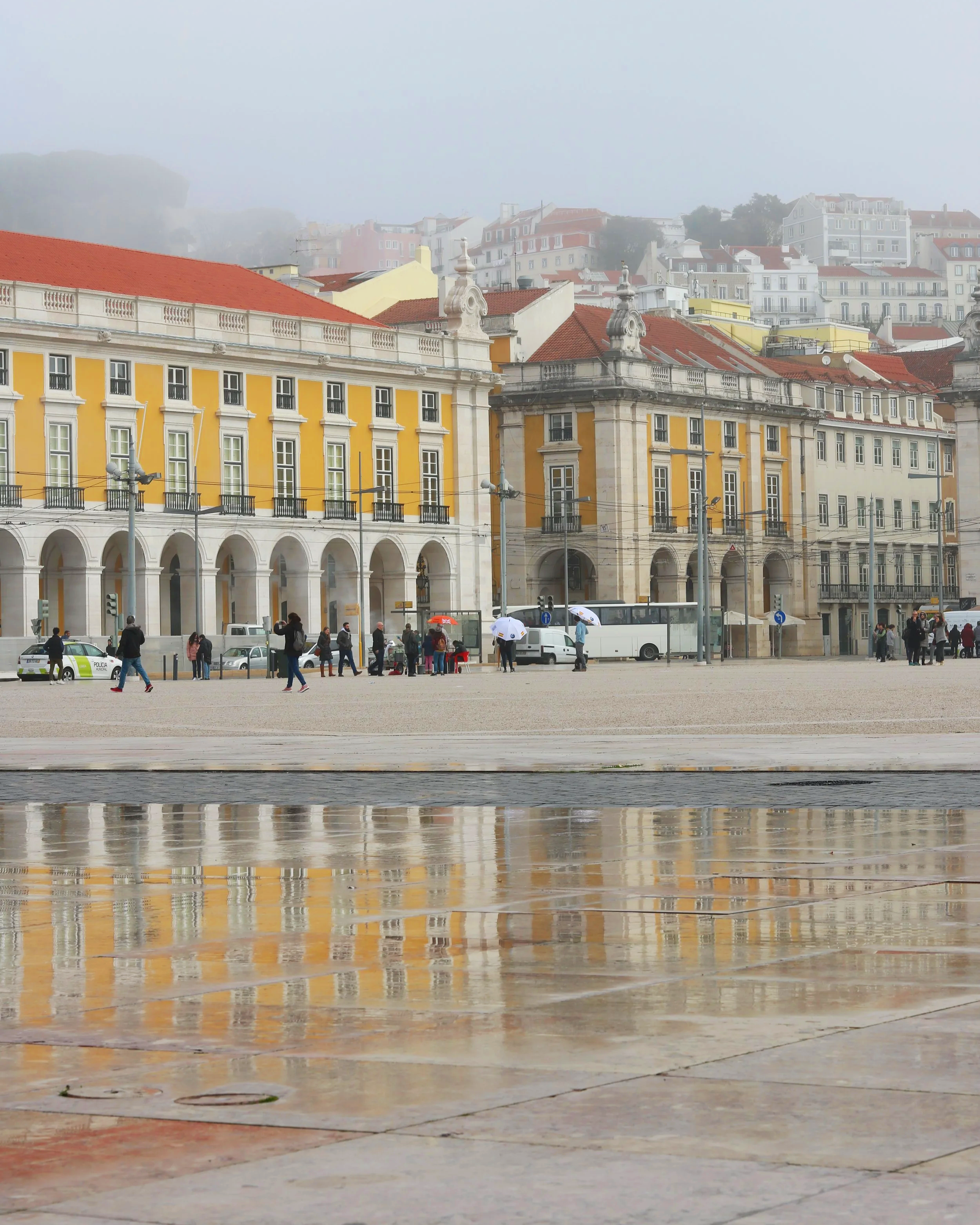 Cityscape of buildings with red roofs, some with ornate architectural details, and a fountain in the foreground reflecting in water, with a foggy background of hilly terrain and additional buildings.