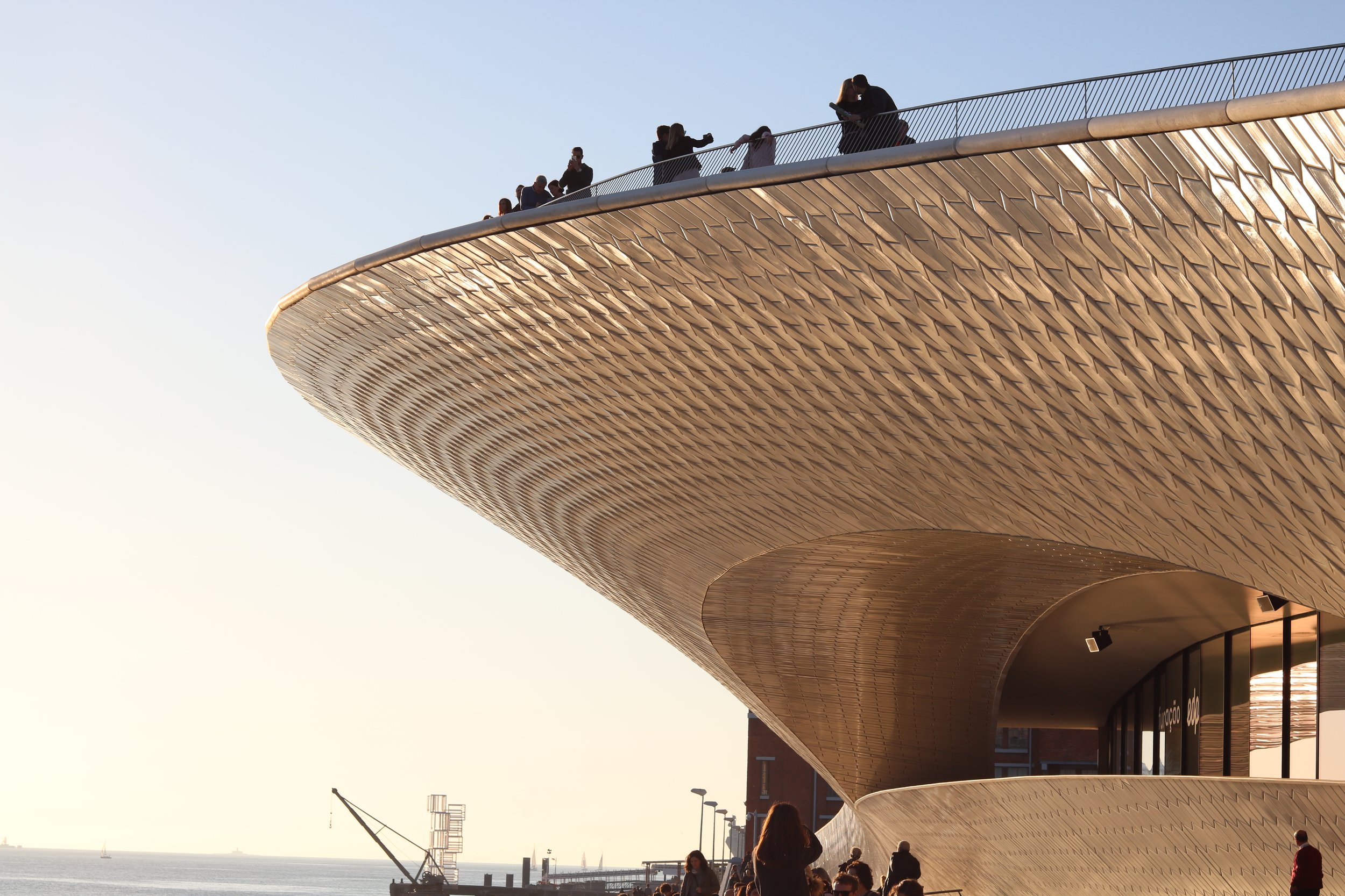 People walking and sitting on a modern, curved building with a textured gold exterior, overlooking a body of water with boats in the distance during daytime.