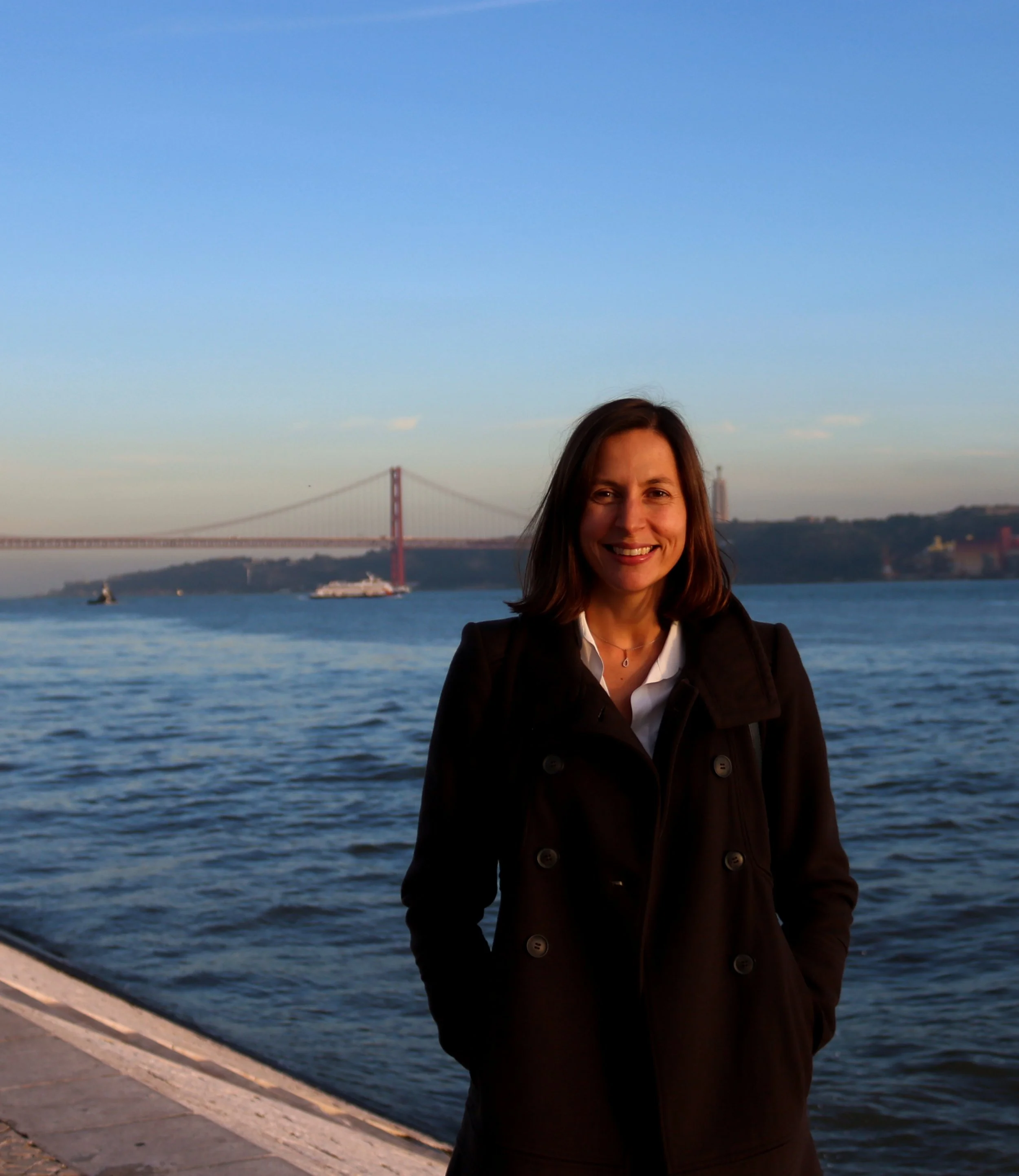 A smiling woman with shoulder-length brown hair stands near a body of water with a bridge in the background during sunset.