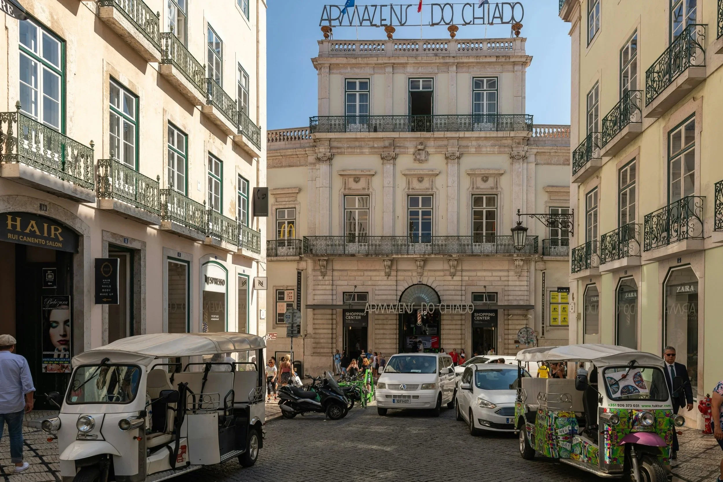 A busy city street with parked cars, motorcycles, and pedestrians. Surrounding buildings have balconies and retail shops, with a large historic building in the background, and a sign in Portuguese that reads 'Arrmazens Do Chiado'.