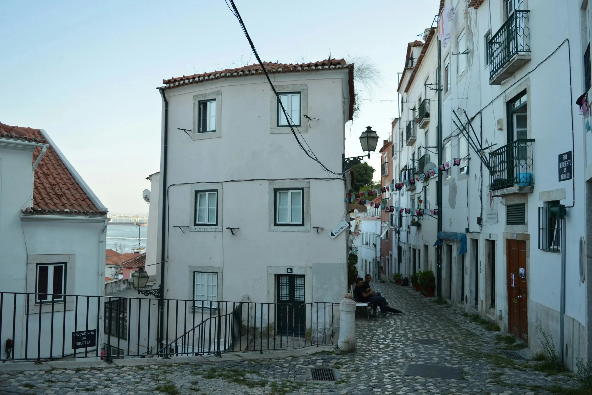 A narrow street with cobblestone pavement, white buildings with red-tiled roofs, hanging laundry, and a street sign that reads 'Rua Norberto de Araújo'. Two people are sitting on a bench, and a street lamp is visible. Wires are strung across the buildings.