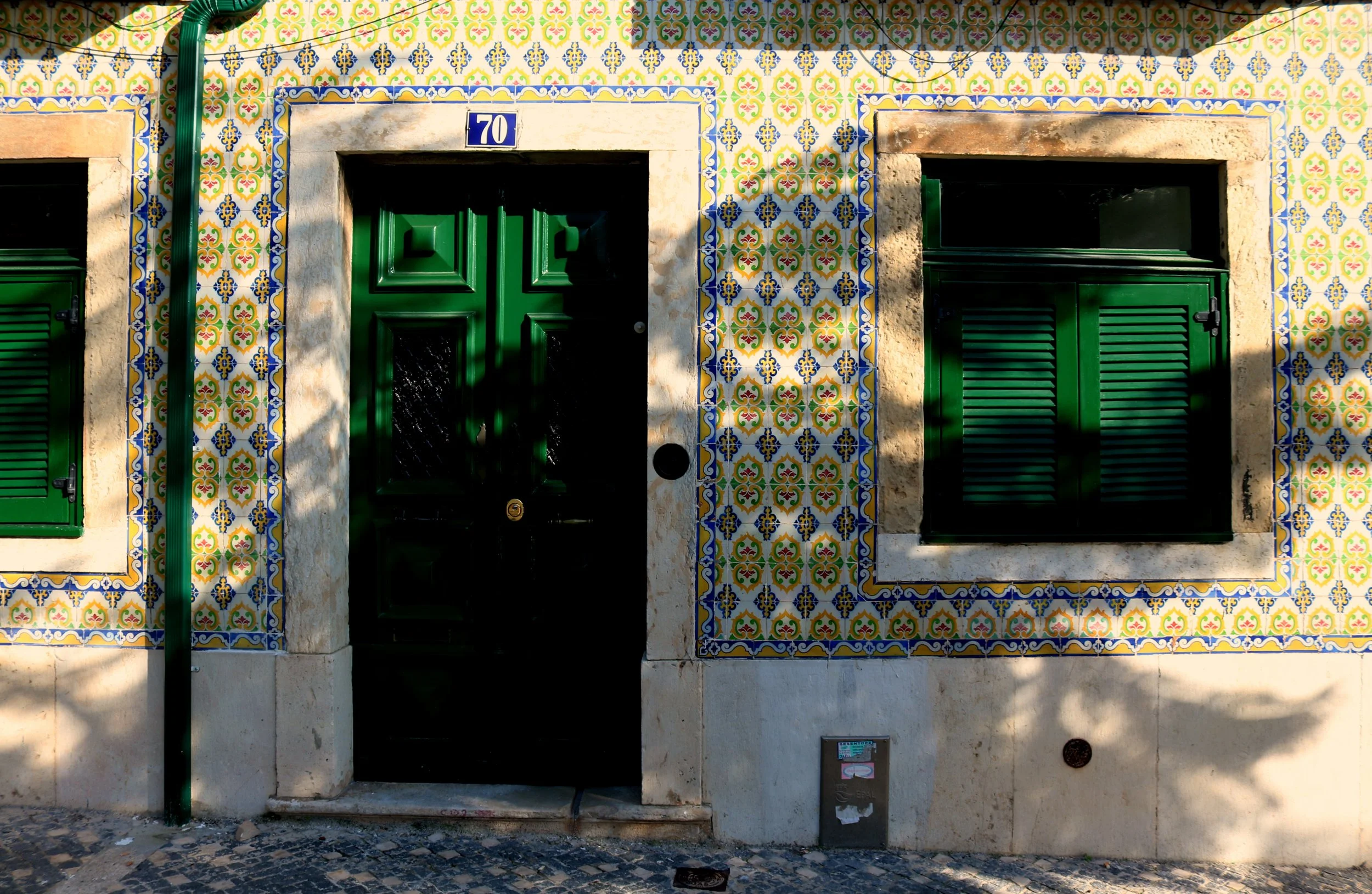 Facade of a building with colorful decorative tiles in yellow, green, blue, and red, featuring a black door with a green frame and ventilation shutters, a blue house number