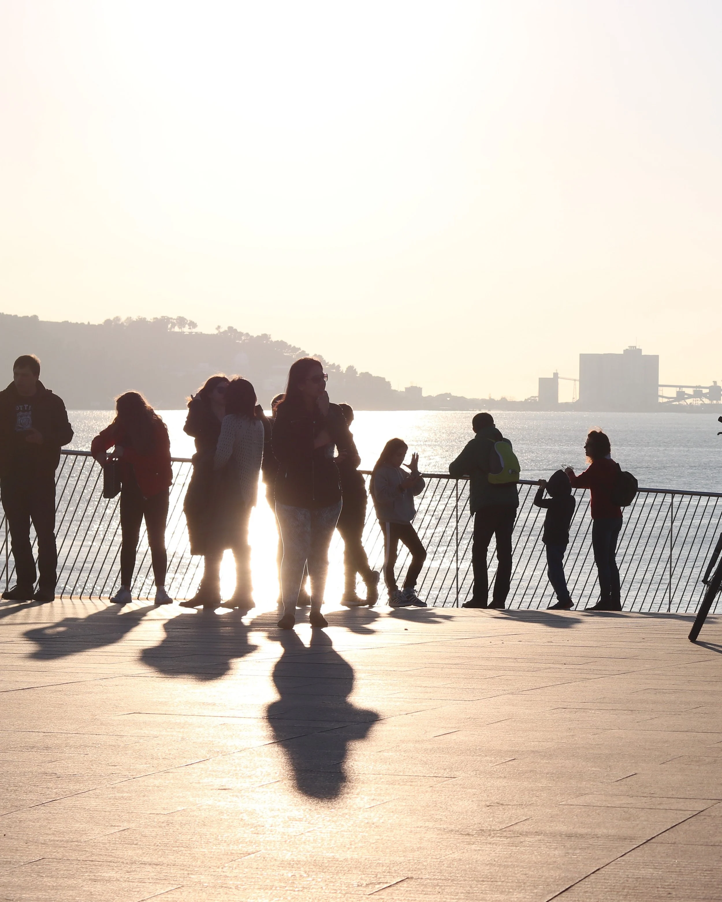 People walking and standing on a waterfront promenade during sunset with visible reflections and shadows on the wooden surface.