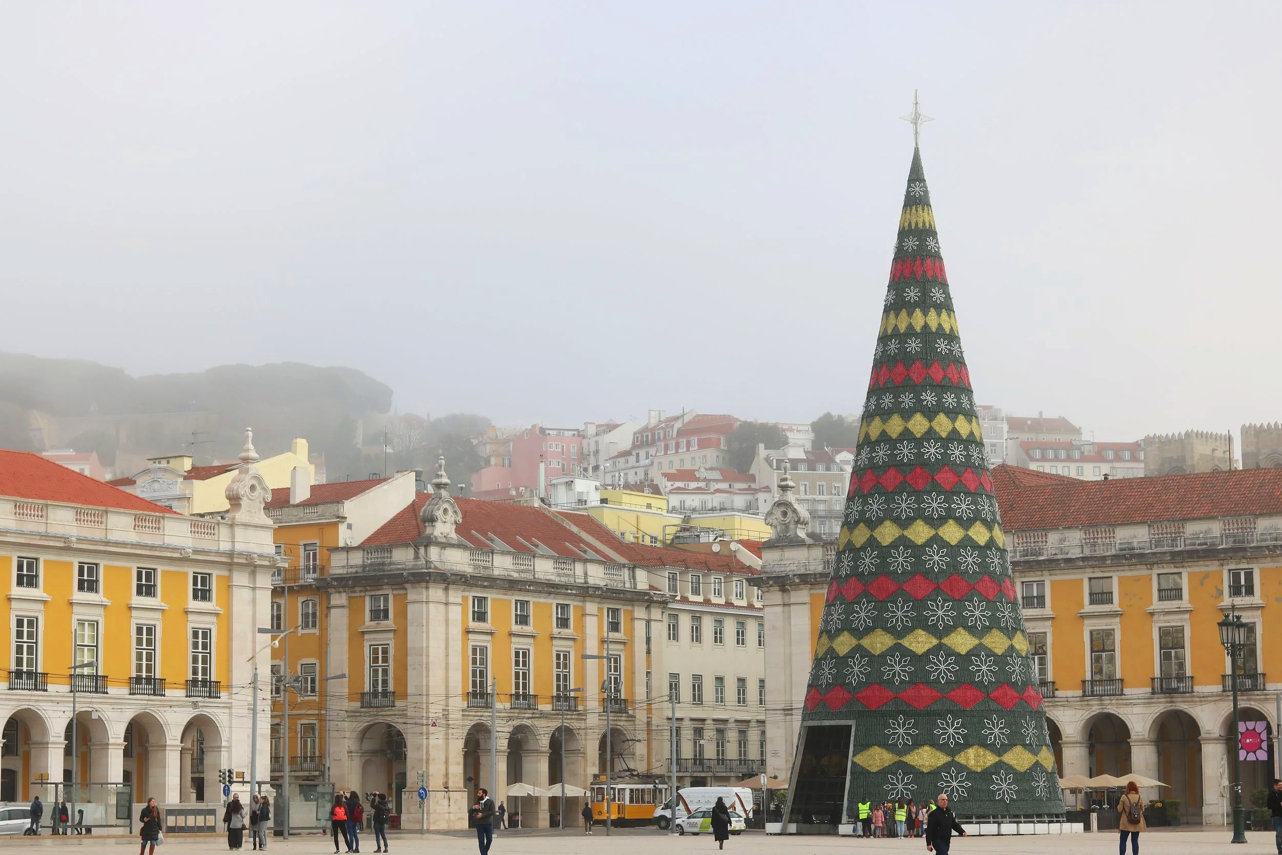 A large cone-shaped Christmas tree with colorful patterns, situated in a city square with historic European-style buildings with yellow facades and red roofs in the background. People are walking around the square.
