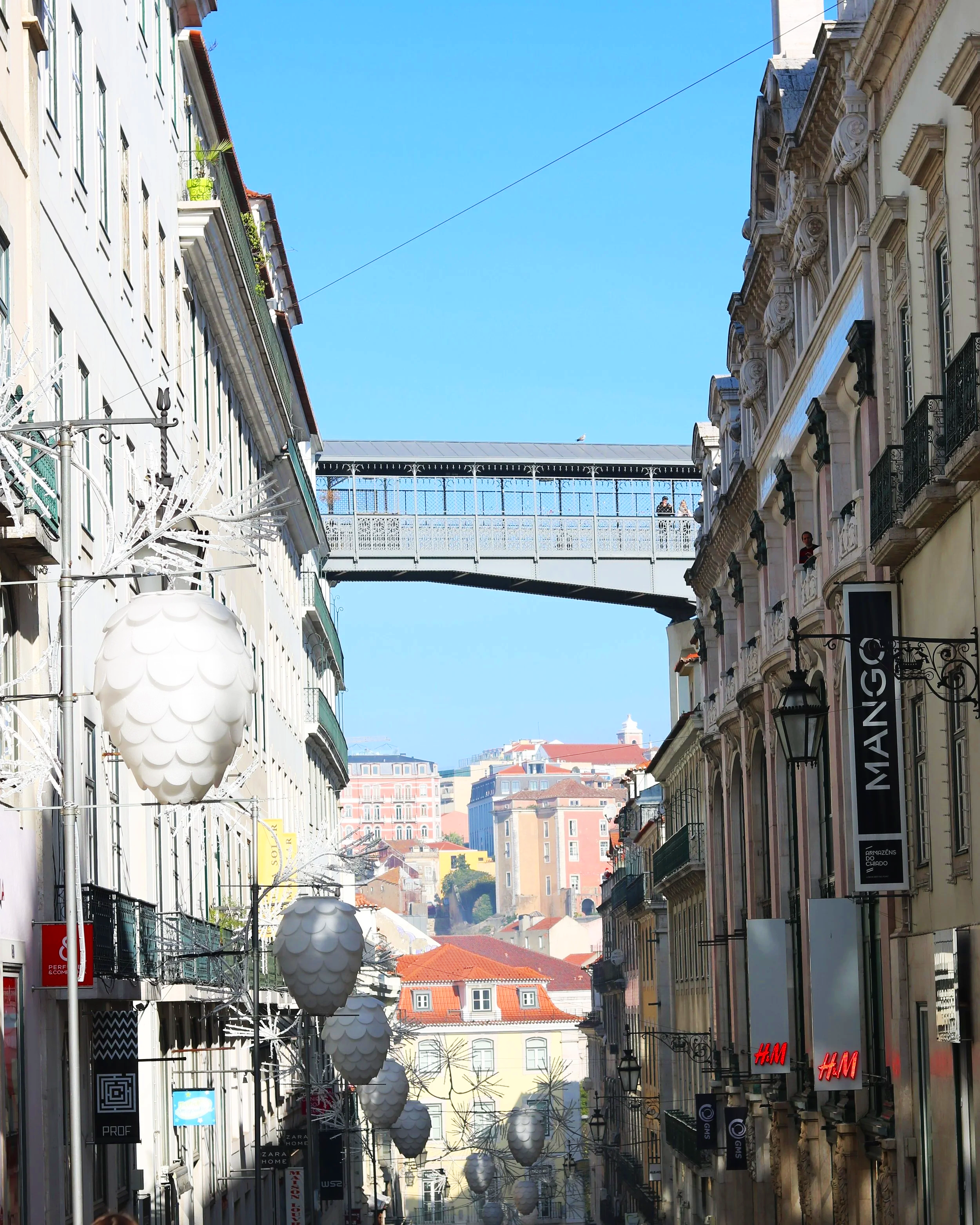 A street with shops and historic buildings on either side, decorated with large white pendant lights hanging overhead, and a background view of city hills and a blue sky.