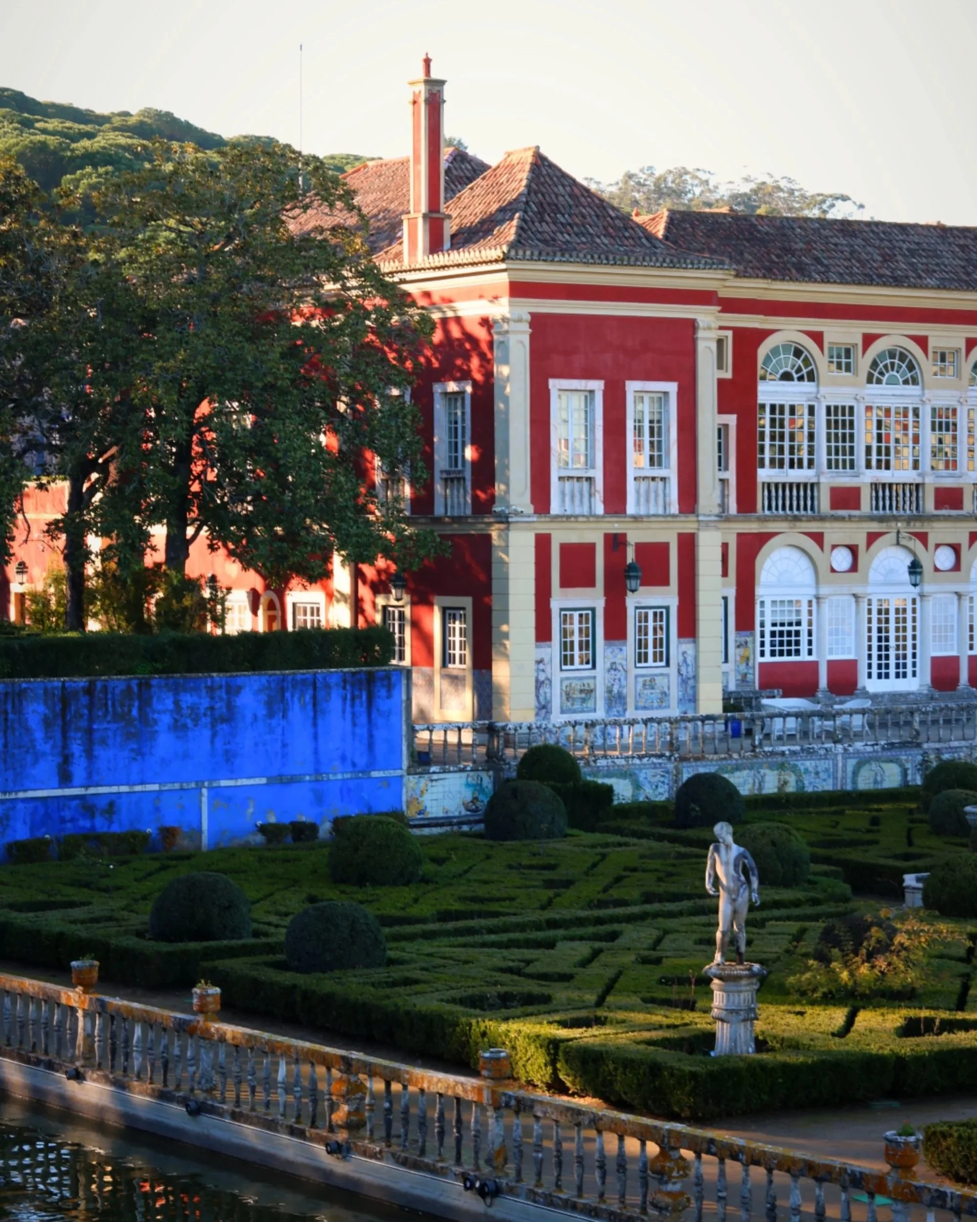 A large, historic building with a red and beige facade, ornate architectural details, and tall arched windows, set behind a formal garden with trimmed hedges, statues, and a waterway in the foreground.