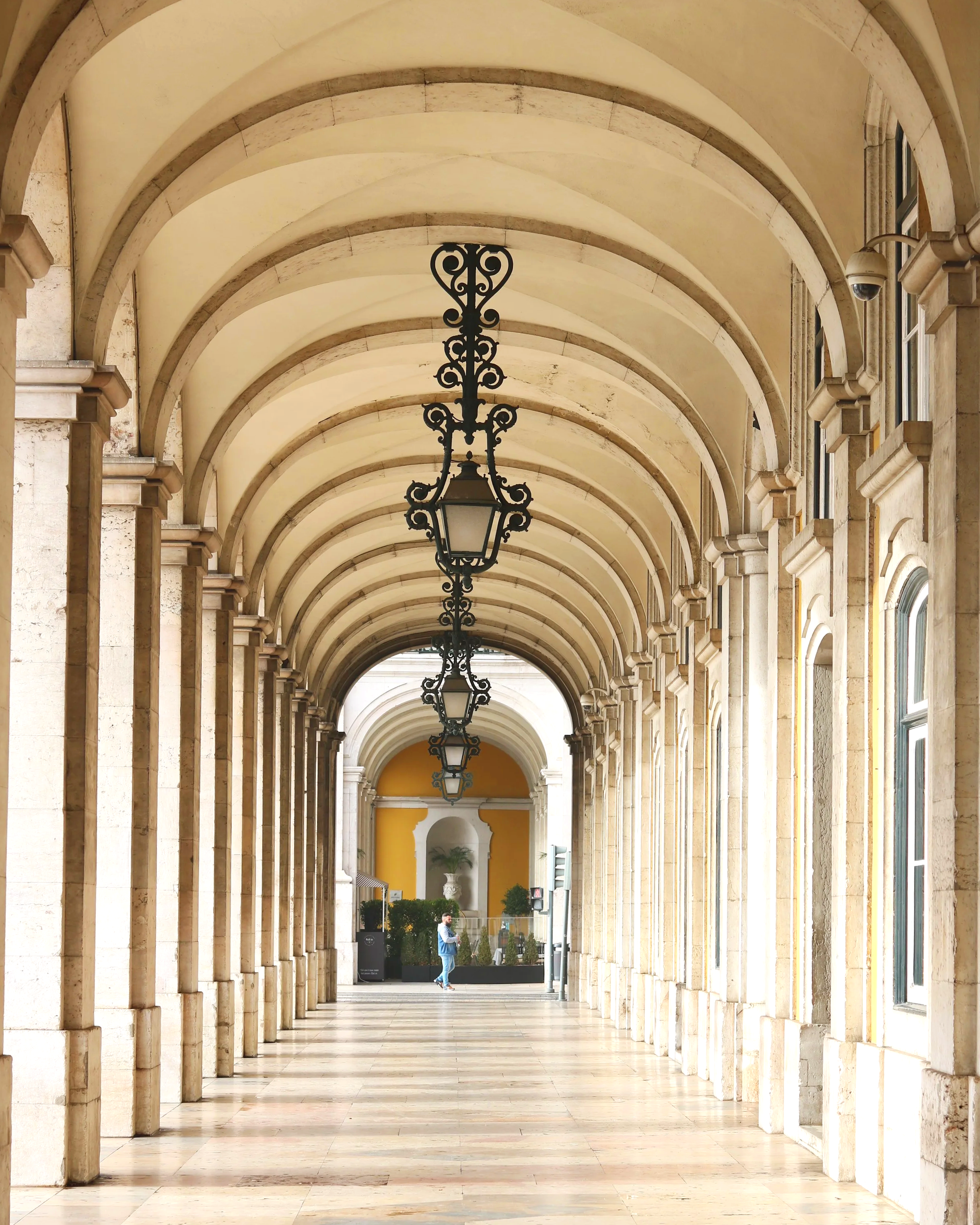 A person in a blue shirt and jeans standing in a long covered walkway with arched ceiling and hanging lanterns, looking at their phone.