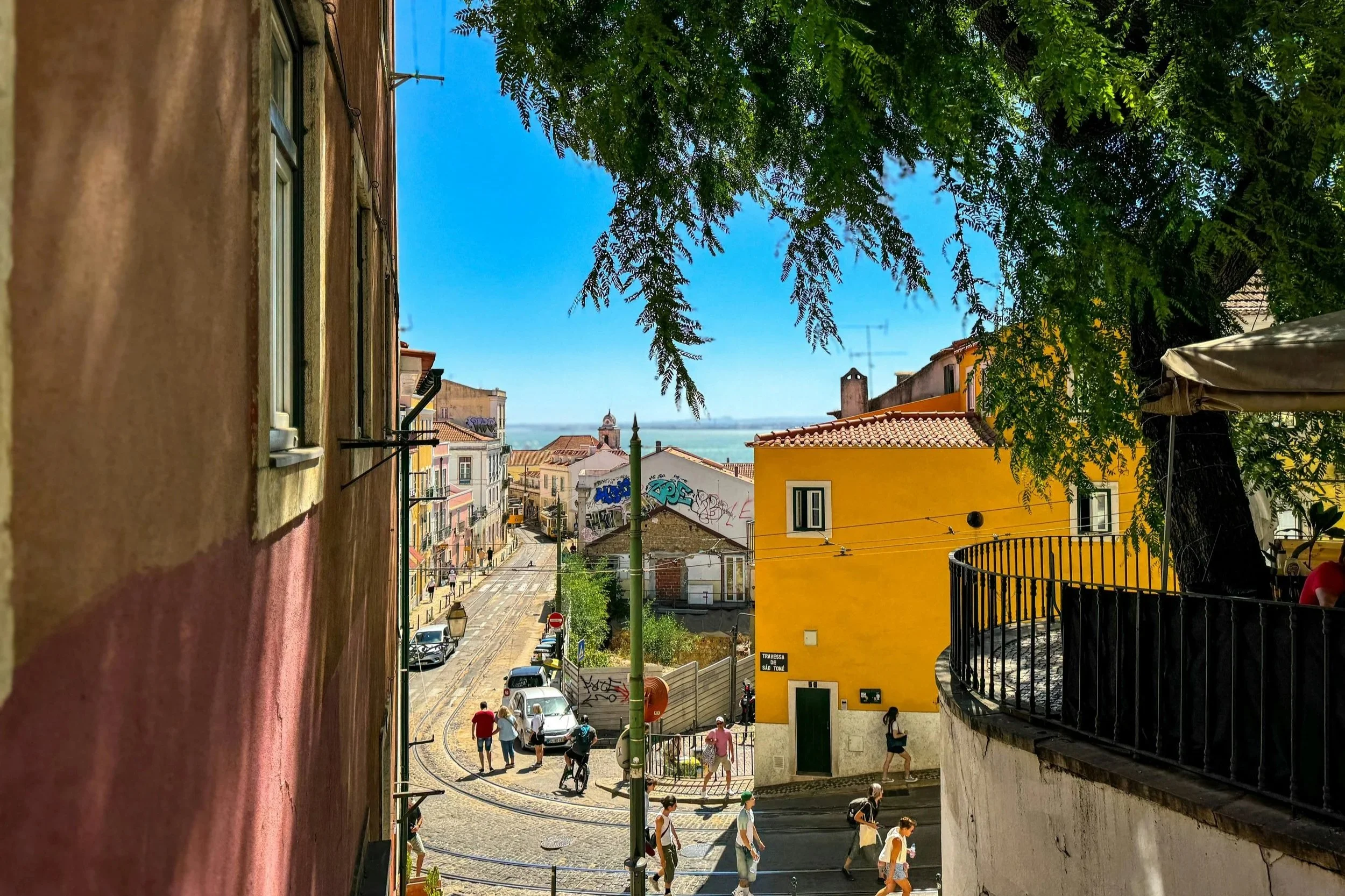 View from a narrow alley down a hill towards a street filled with pedestrians, parked cars, and tram tracks, with colorful buildings, trees, and a clear blue sky in the background.