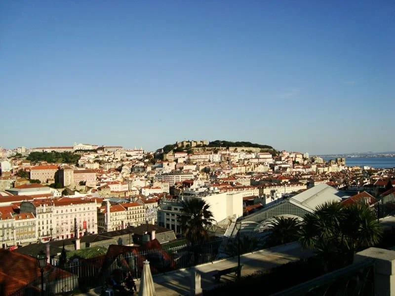 A panoramic view of a European city with a hilltop fortress, colorful buildings, and a waterfront in the distance under a clear blue sky.
