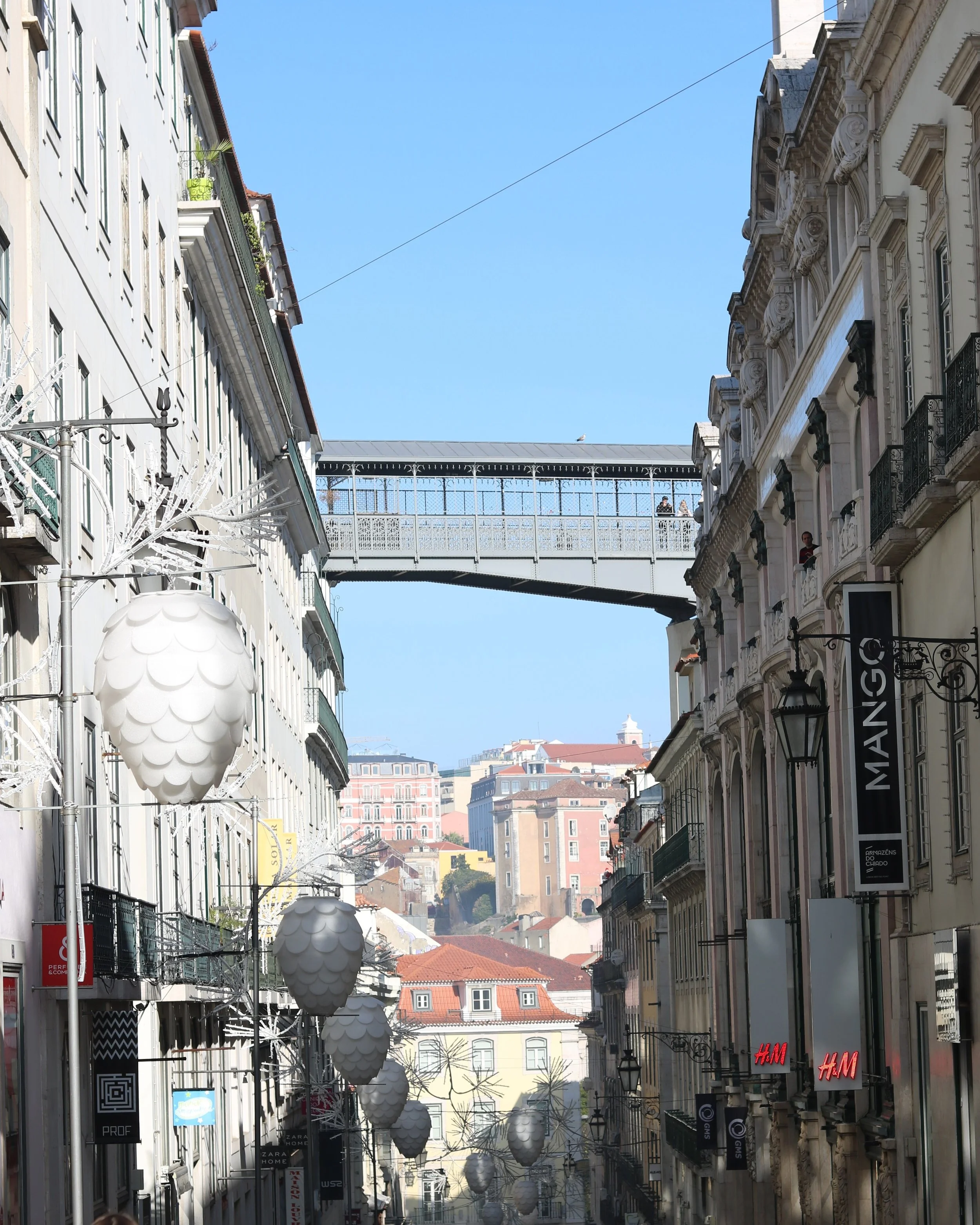 A street scene with buildings lining both sides, decorative street lamps hanging overhead, and a bridge connecting the buildings in the background under a clear blue sky.