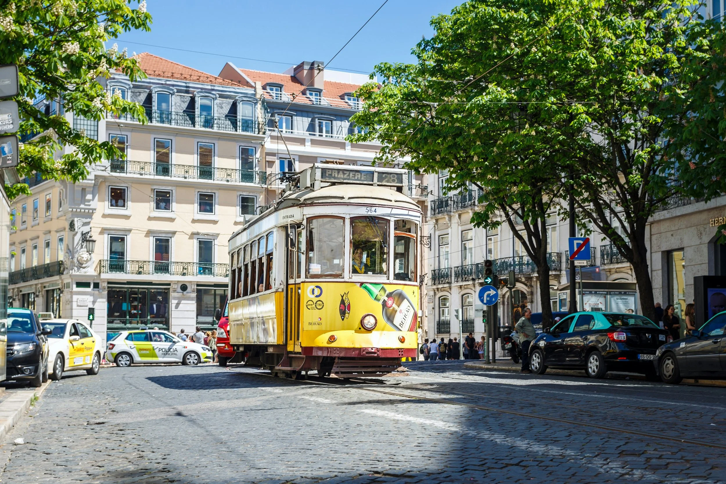 A yellow tram traveling down a city street surrounded by cars, pedestrians, and green leafy trees, with buildings in the background under a clear blue sky.