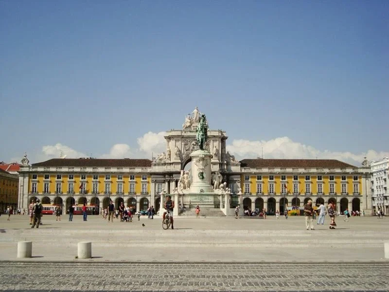 A large historic square with a monument featuring a statue at the center, surrounded by a yellow and white building, with people walking and biking around under a partly cloudy sky.