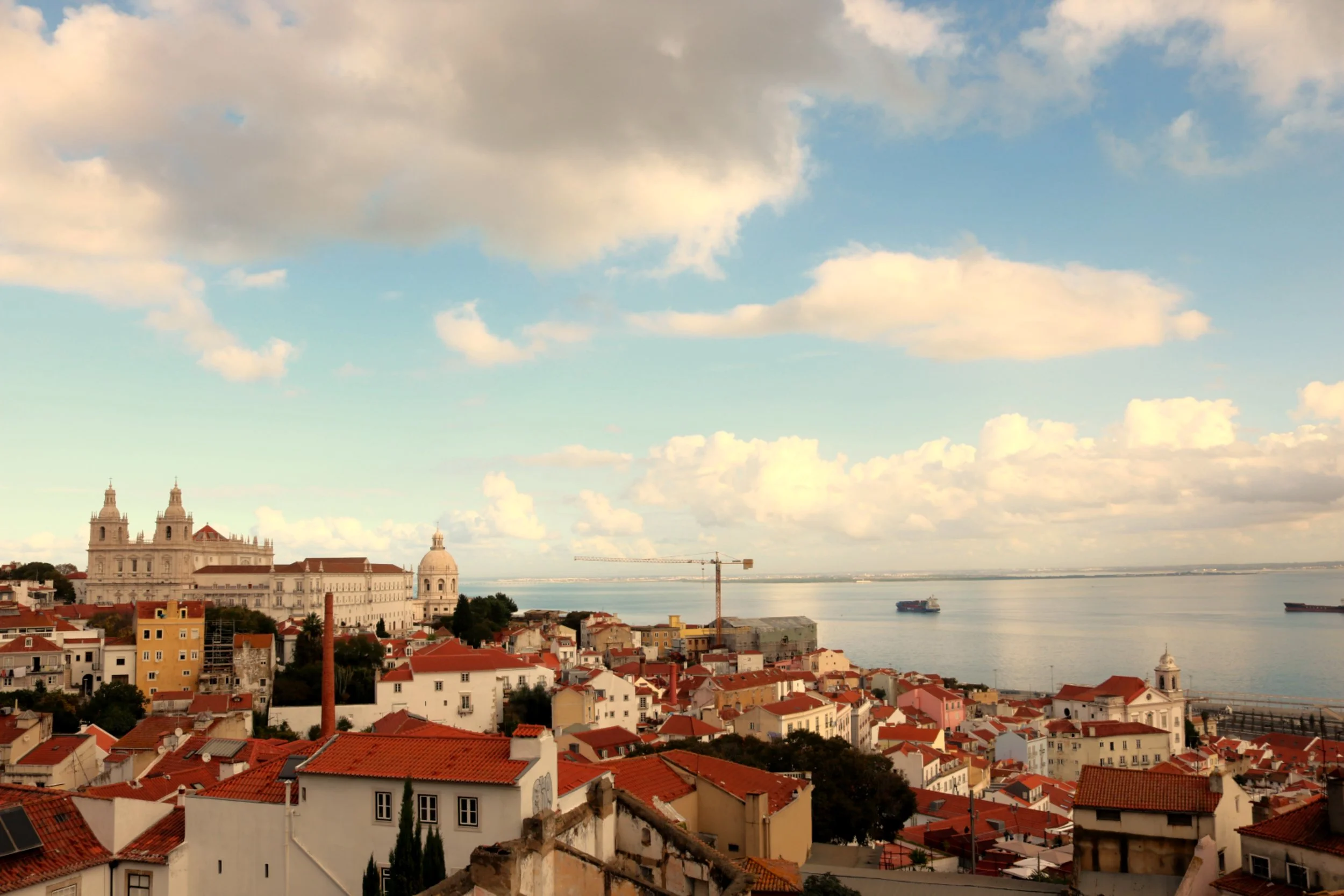 A cityscape with historic white buildings with red-tiled roofs, a large river in the background, and a sky filled with fluffy clouds.