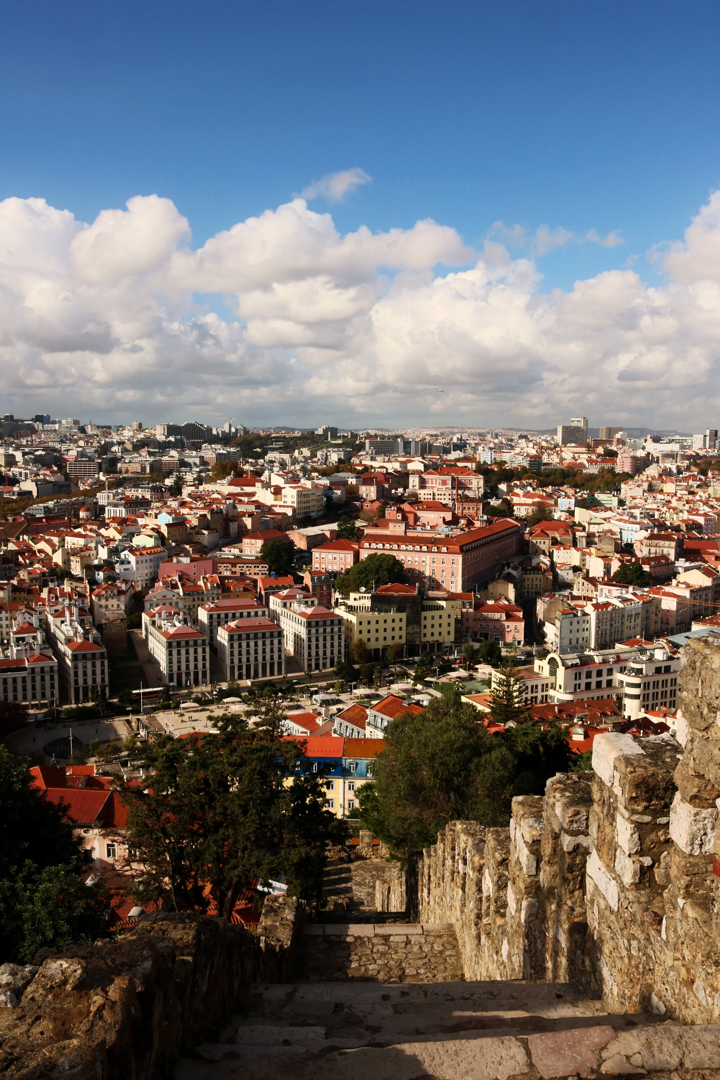 Cityscape of Lisbon, Portugal viewed from a historic site with stone stairs and walls in the foreground, under a partly cloudy sky.