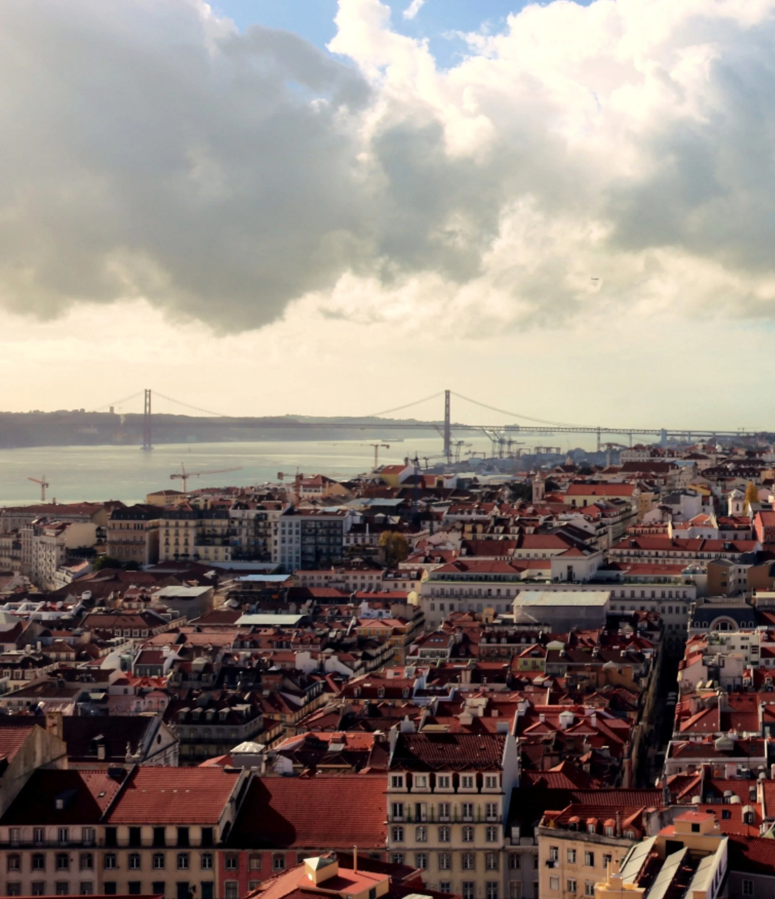 A cityscape with numerous rooftops, a body of water, and a bridge in the distance under a cloudy sky.