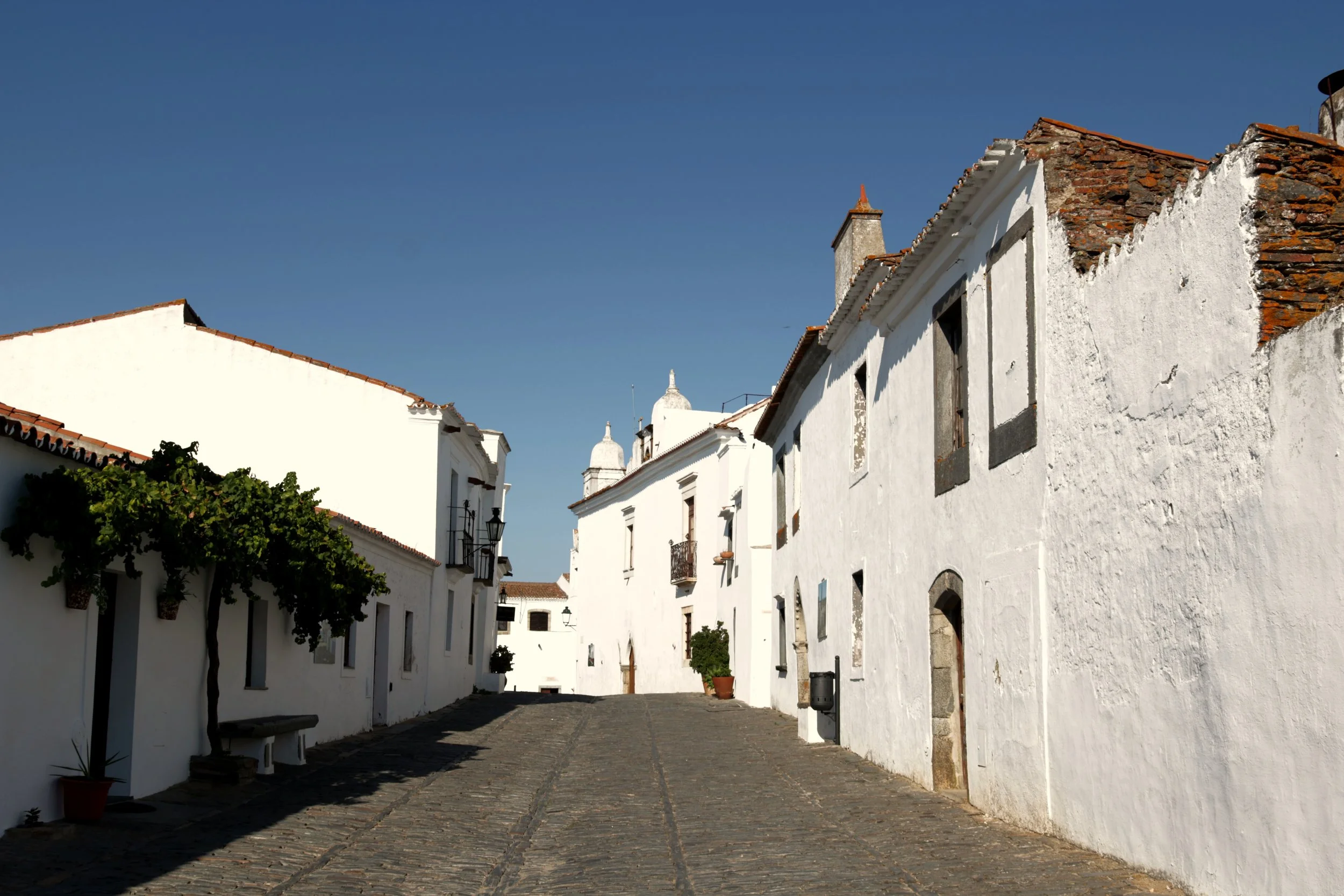 A quiet street lined with white buildings with tiled roofs under a clear blue sky.