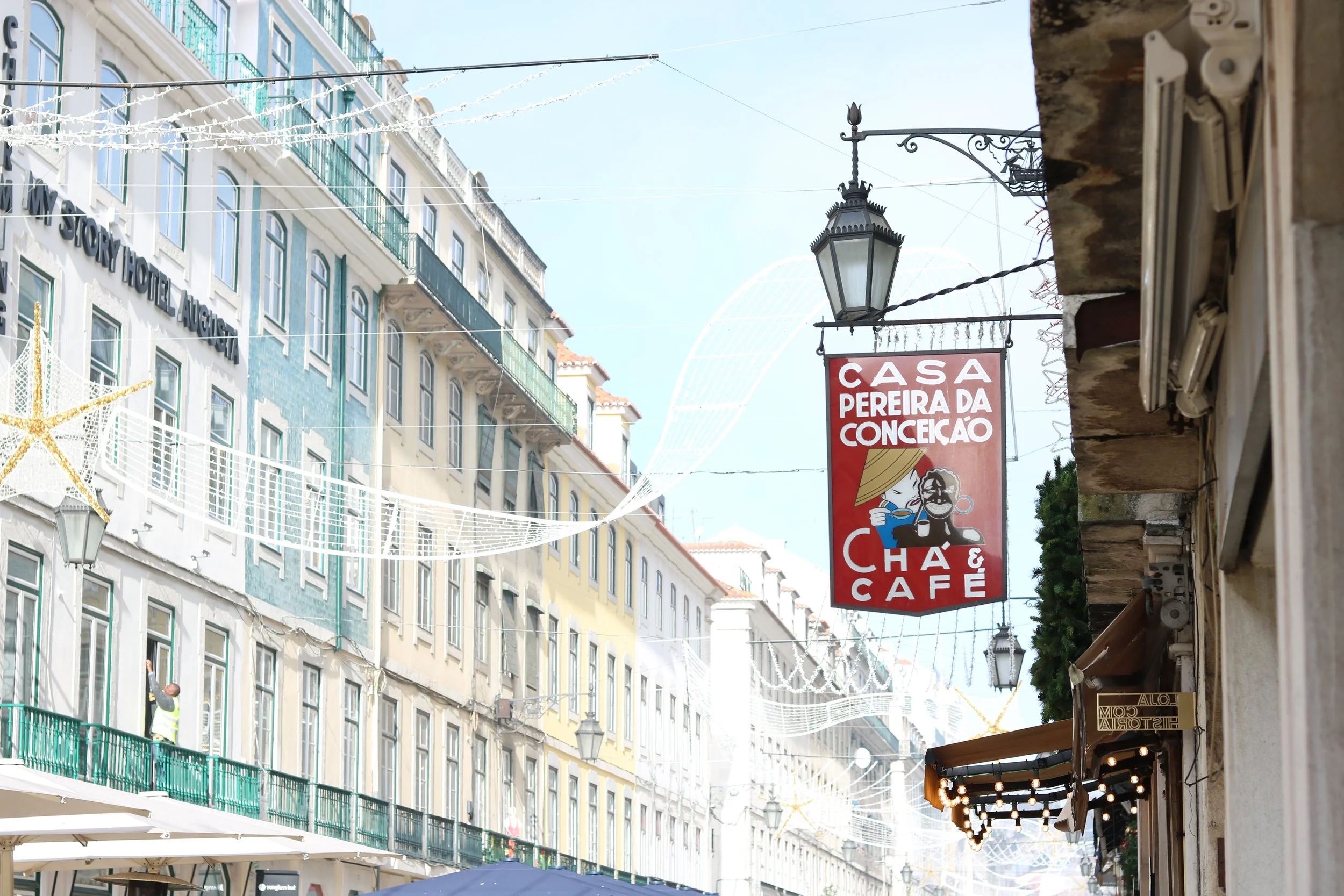 Street scene with colorful buildings, hanging holiday lights, a street lamp, and a red sign for Casa Pereira da Conceição, a café, on a sunny day.