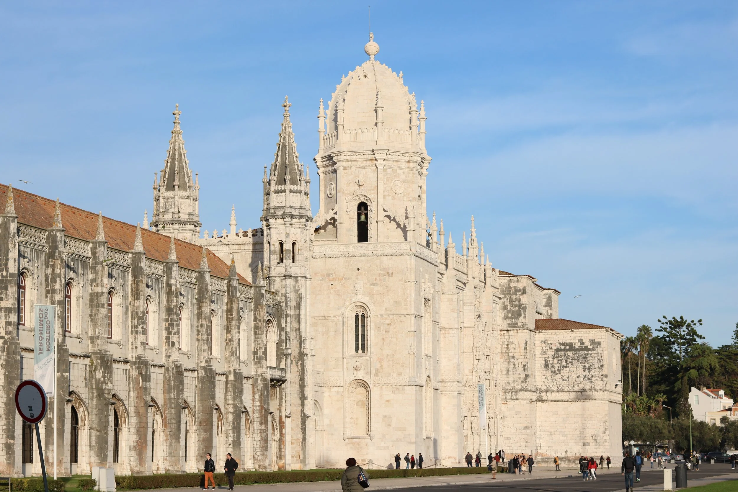 Historical stone building with ornate towers and spires, with people walking and standing in front under a clear blue sky.