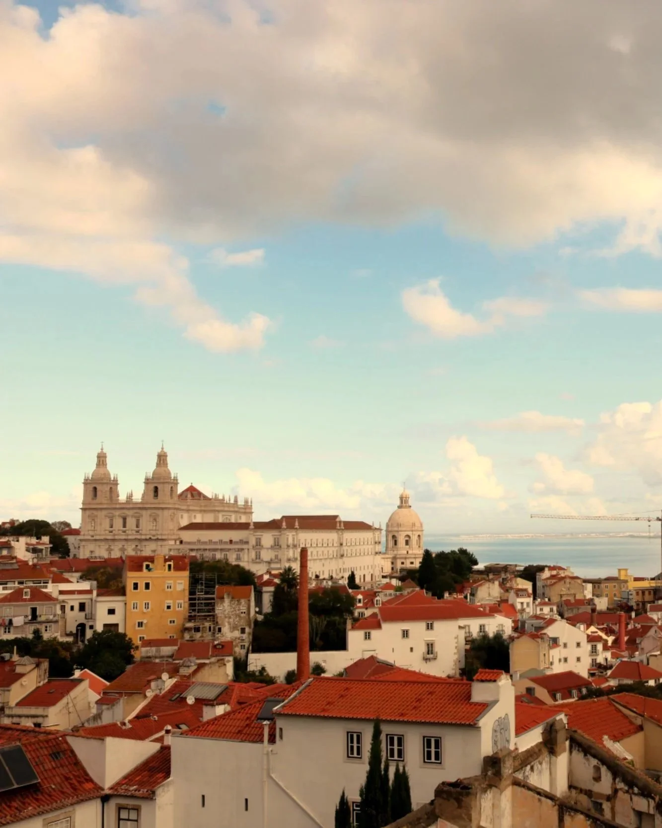 A cityscape with historic buildings and red-tiled roofs under a partly cloudy sky, overlooking a body of water in the distance.