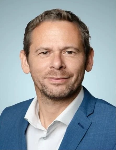 Headshot of Andres Moll, co-founder of Career Circle Luxembourg, with light brown hair and a beard, wearing a blue suit and white shirt, smiling at the camera against a light blue background.