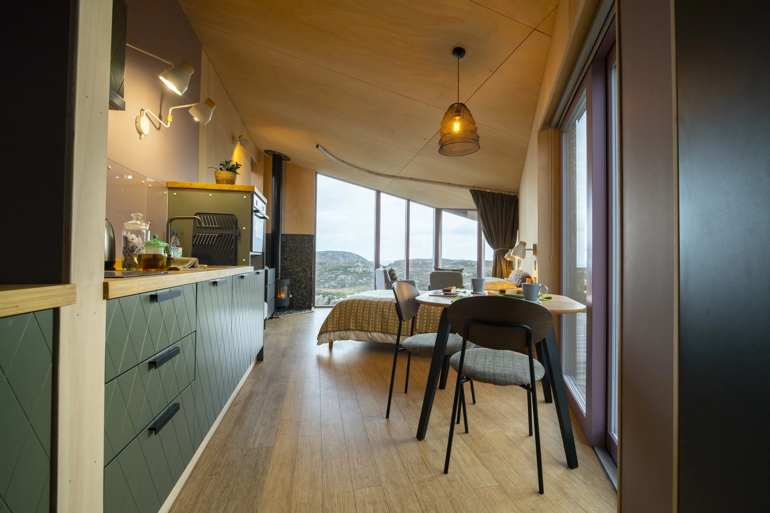 Interior of a modern living space with wooden ceiling, large windows showing a scenic landscape, green kitchen cabinets, a dining table with chairs, and decorative lighting fixtures.