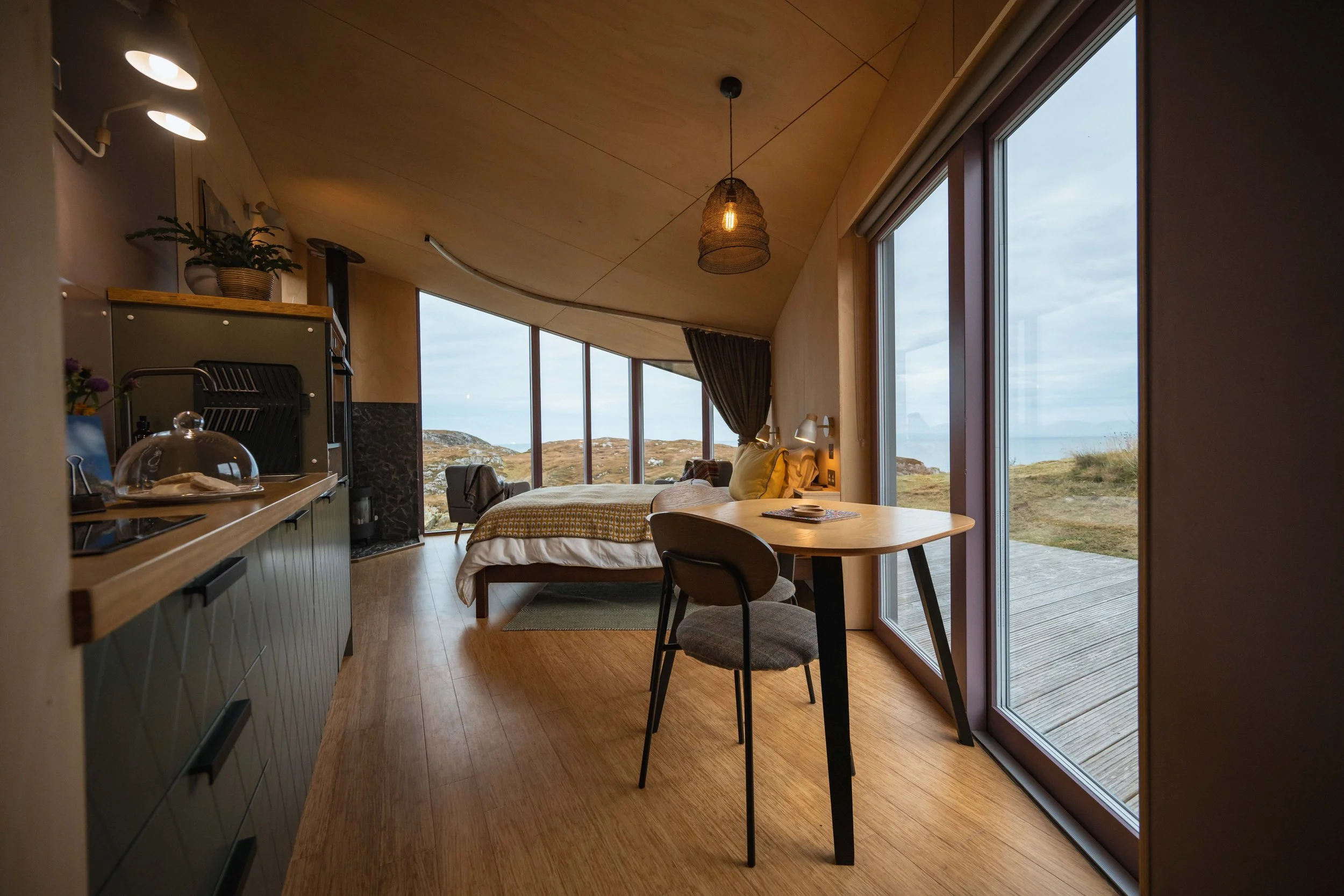Interior of a modern living space with a large view of a landscape through floor-to-ceiling windows, wooden ceiling, small kitchen area with green cabinets, a dining table with chairs, a bed, and hanging pendant lights.