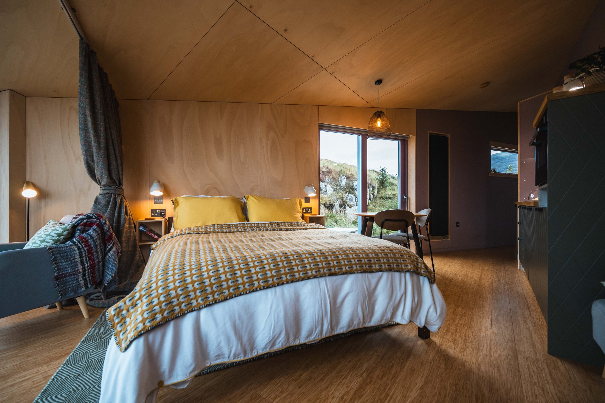 A cozy bedroom with wooden wall panels and ceiling, featuring a large bed with yellow and white bedding, two bedside lamps, a plaid blanket, a window showing outdoor scenery, and a small table with chairs.