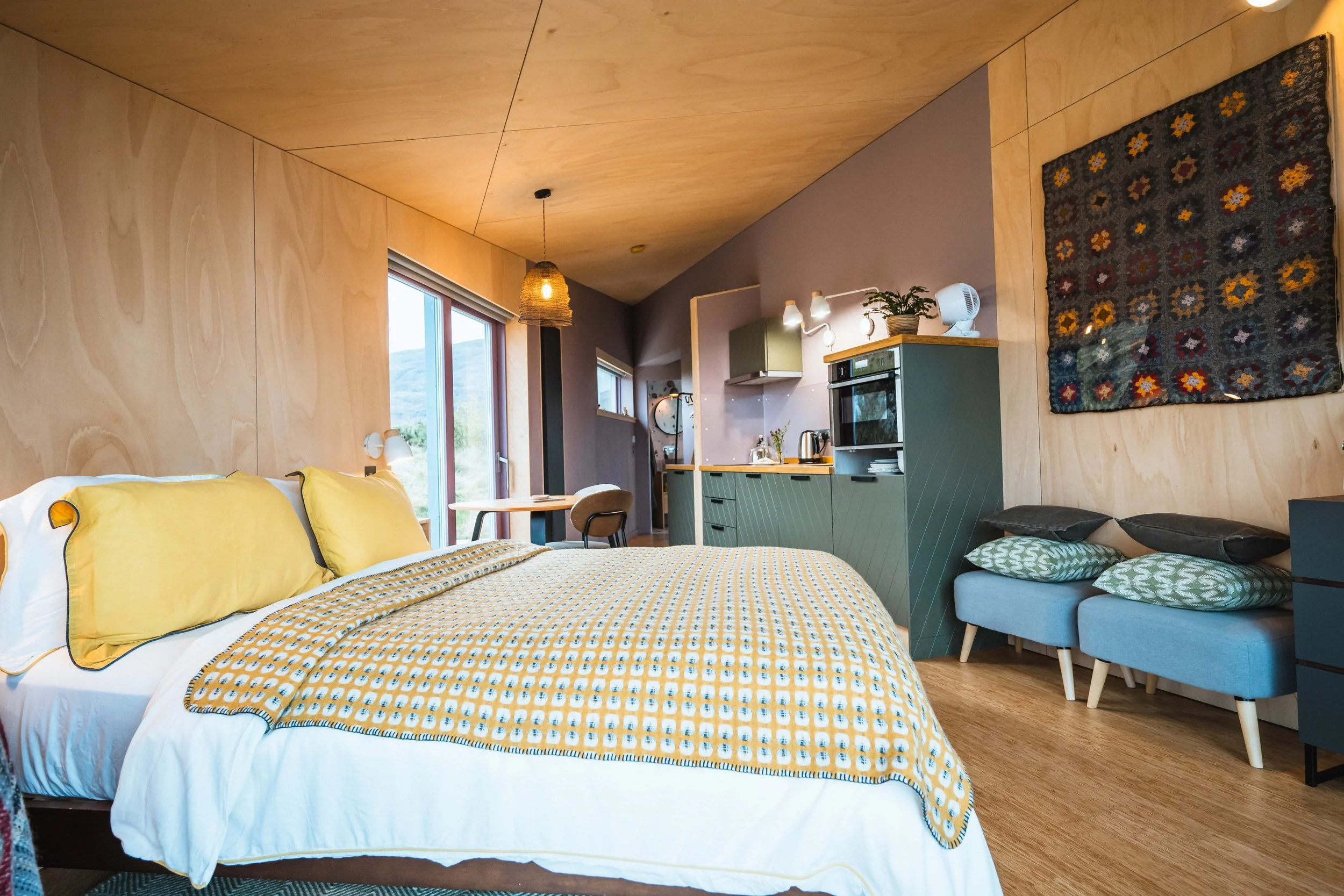 A cozy bedroom with a bed featuring yellow pillows and a yellow and white patterned blanket. The room has wood-paneled walls and ceiling, a window with a view outside, and a small sitting area with a light blue armchair. The kitchen area includes a microwave, kettle, and a wall-mounted cabinet. There is a decorative textile hanging on the wall and a woven pendant light hanging from the ceiling.