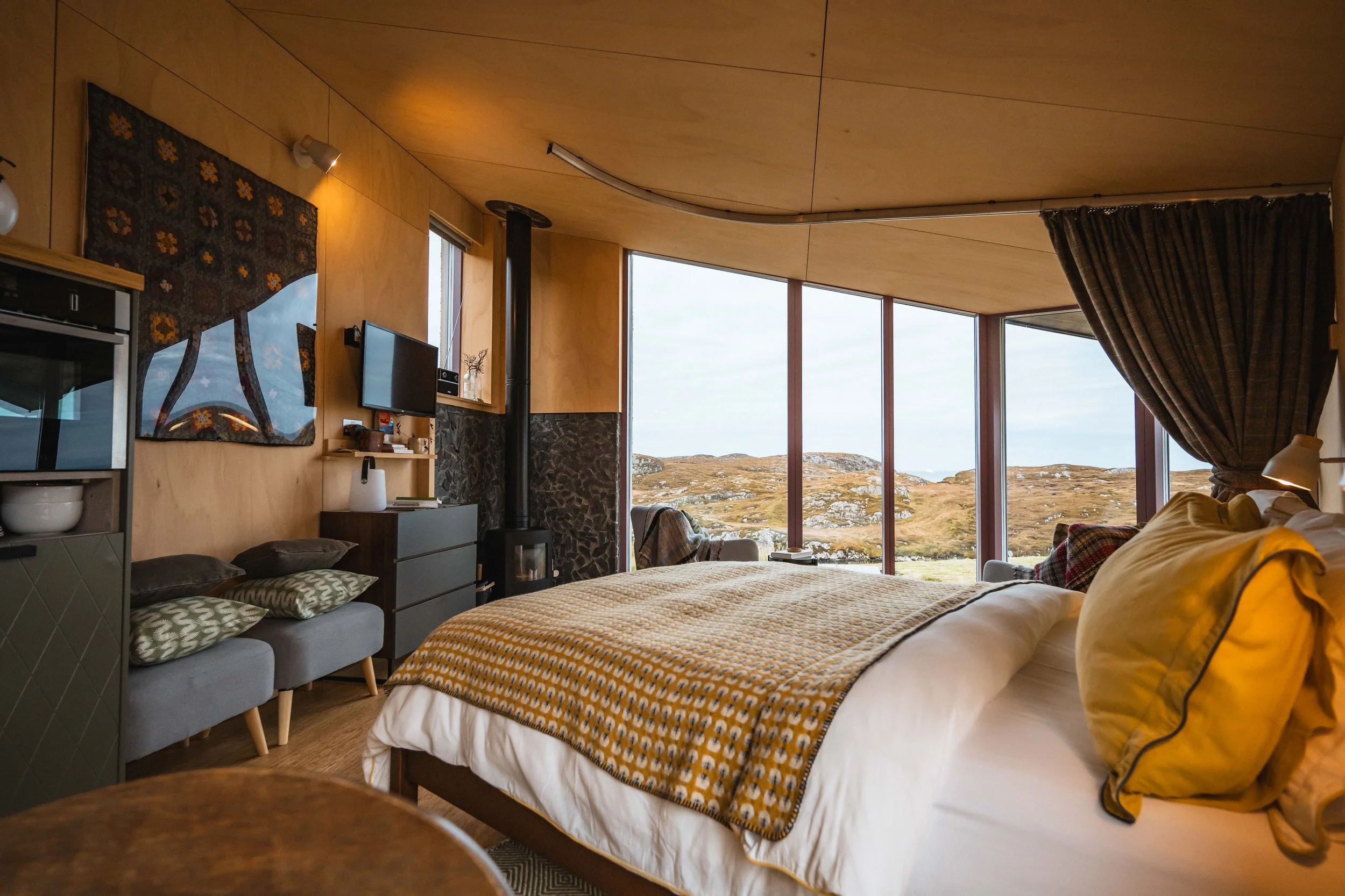 Interior of a cozy bedroom with large floor-to-ceiling windows showing a scenic landscape of grassy hills and rocky terrain outside. The room features a bed with a yellow and white patterned blanket, a small gray sofa with patterned pillows, a flat-screen TV on the wall, and a wood-burning stove with a chimney. Curtains are partially drawn on one side of the windows.