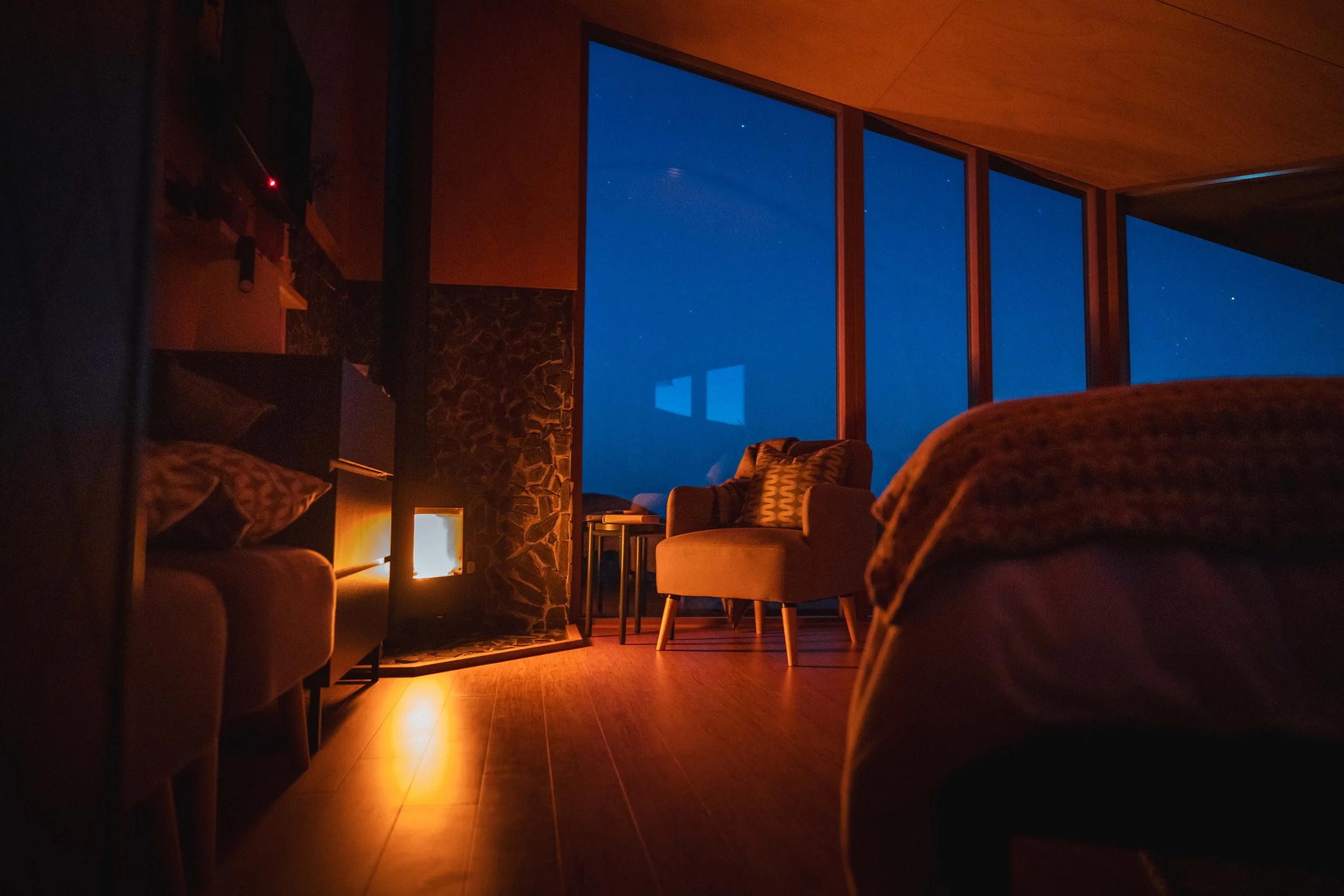 Interior of a dimly lit living room at night with large windows showing a dark sky with visible stars, a cozy seating area with chairs, a stone fireplace, and warm lighting.