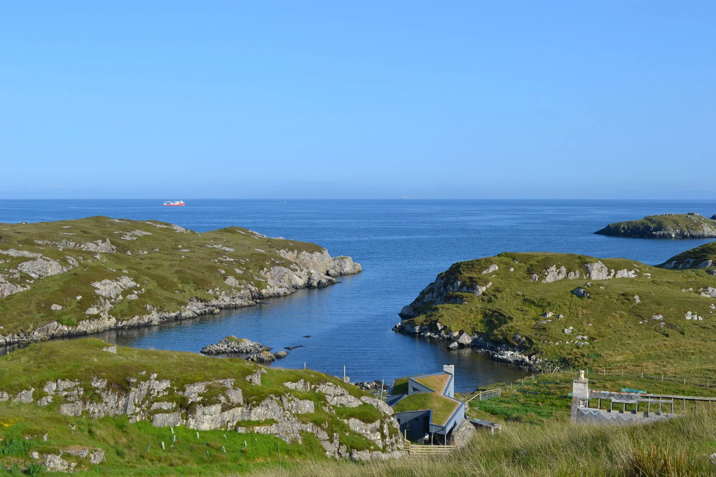 Coastal view with green hills, rocky formations, and small houses overlooking the sea. A boat is seen on the water under a clear blue sky.