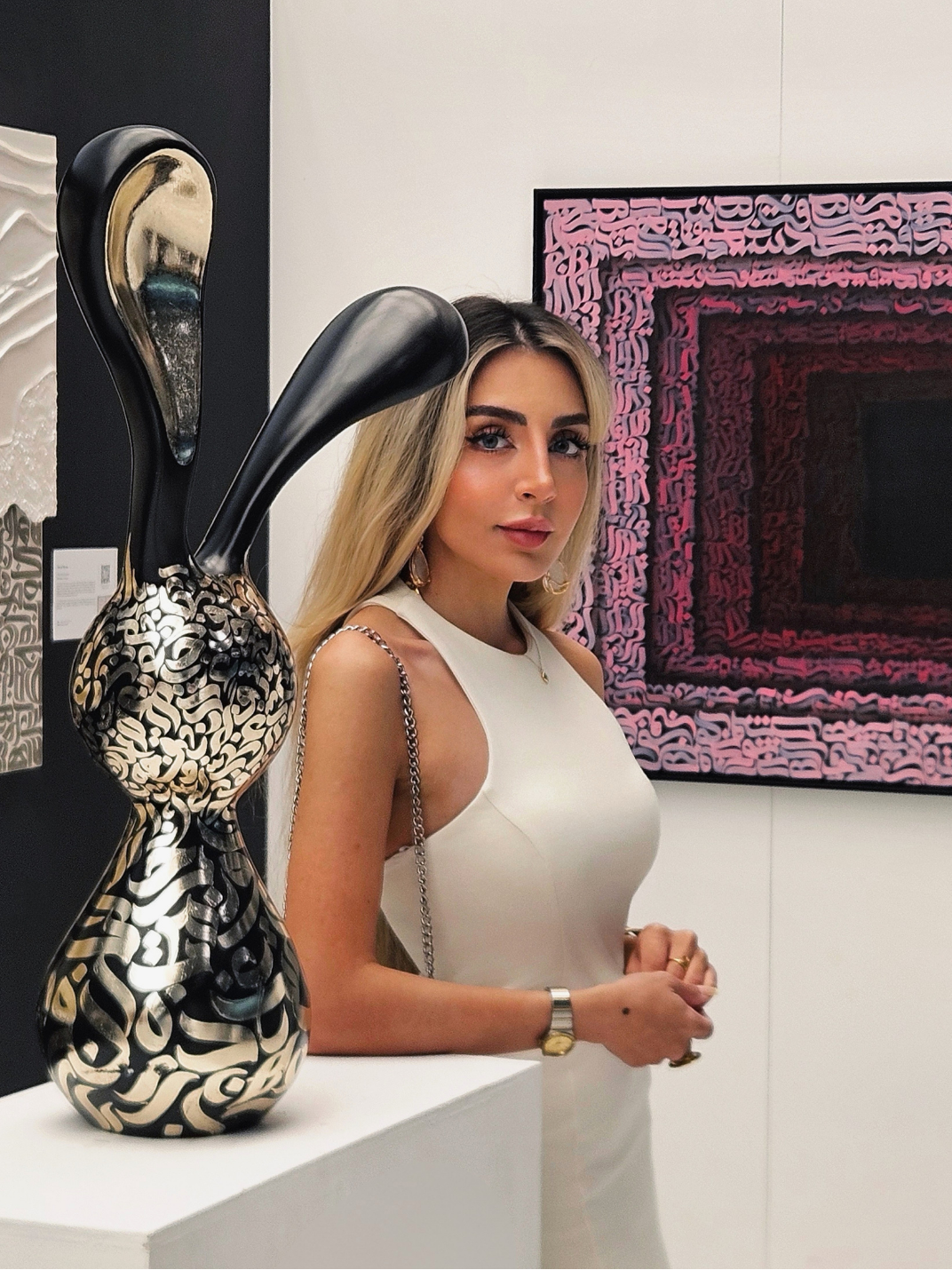 Artist Shirin Roshanaie standing in an art gallery, with her "Nouran" sculpture and colorful Persian and Arabic Calligraphy artwork in the background. ShisArt Studio