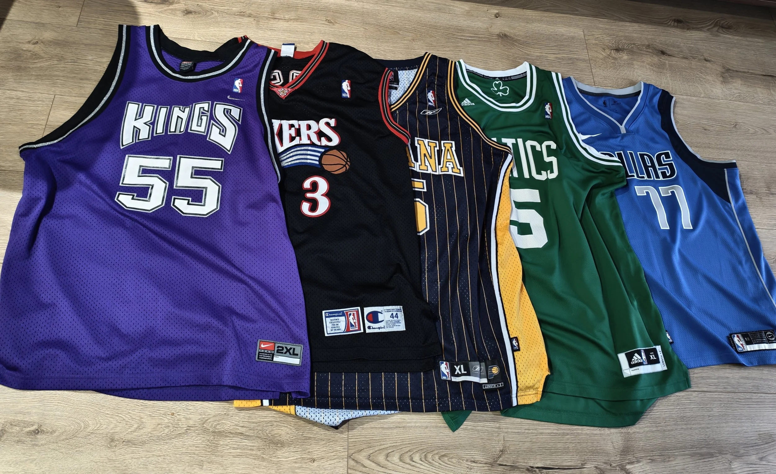 A collection of five NBA basketball jerseys laid out on a wooden floor. The jerseys are purple, black, dark blue, green, and blue, each representing different NBA teams.