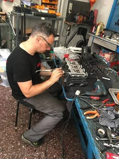 A man working on a car engine in a workshop, sitting at a workbench with tools and parts around him.