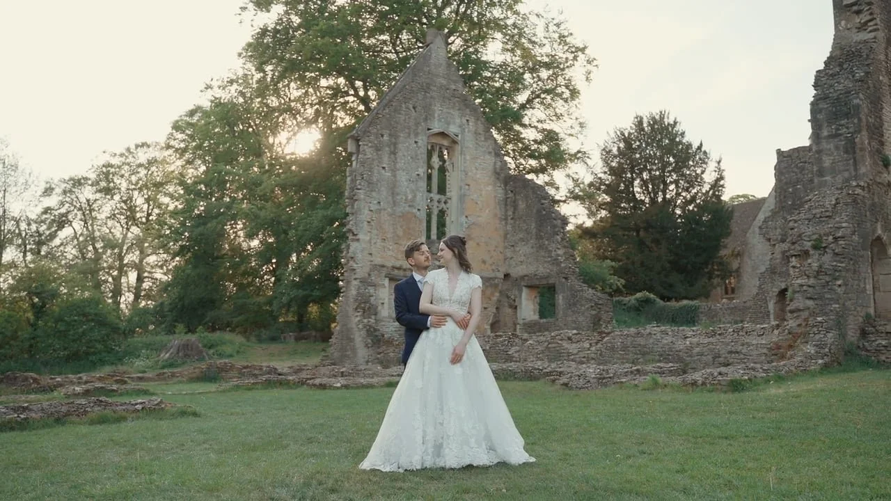 Bride and groom standing together outdoors in front of a partially ruined stone building with trees, during sunset.
