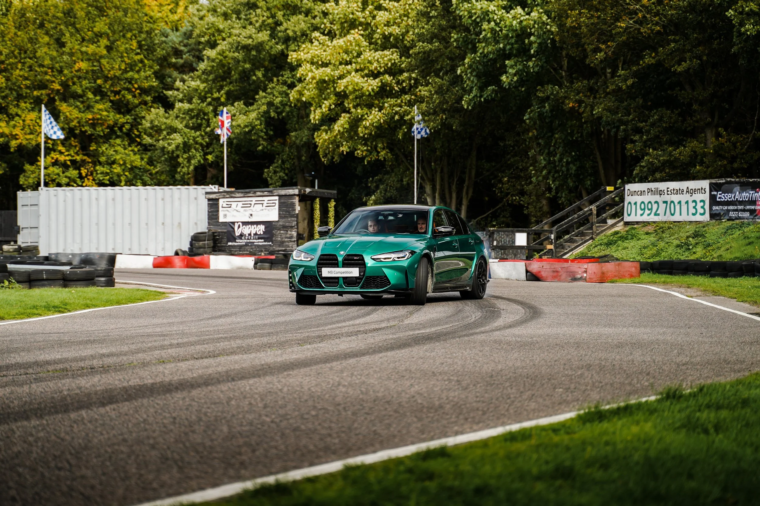 A green modern hatchback car on a race track with a driver and a passenger inside. The background shows trees and advertisements, with flags of various designs flying above the track.