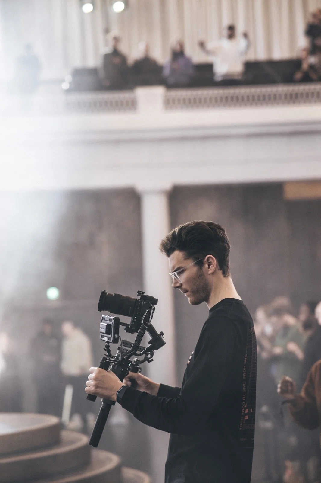 Young man with glasses and dark hair operating a professional camera on a stabilizer in a large indoor space with people in the background.