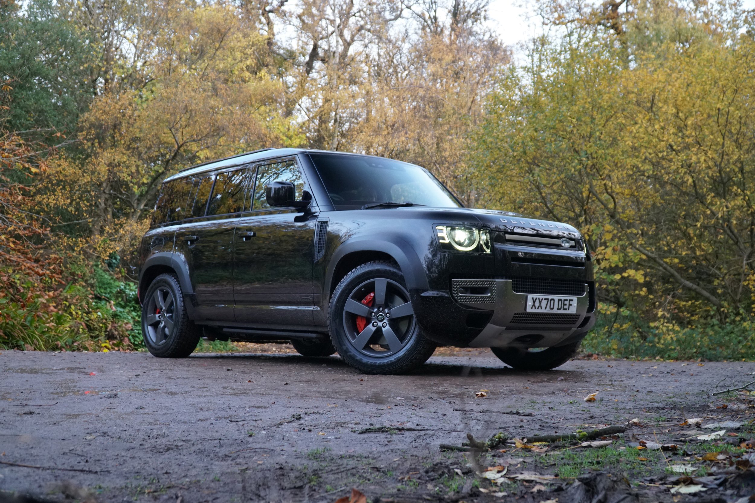 A black Land Rover Defender parked on a dirt road surrounded by autumn trees with yellow and green leaves.