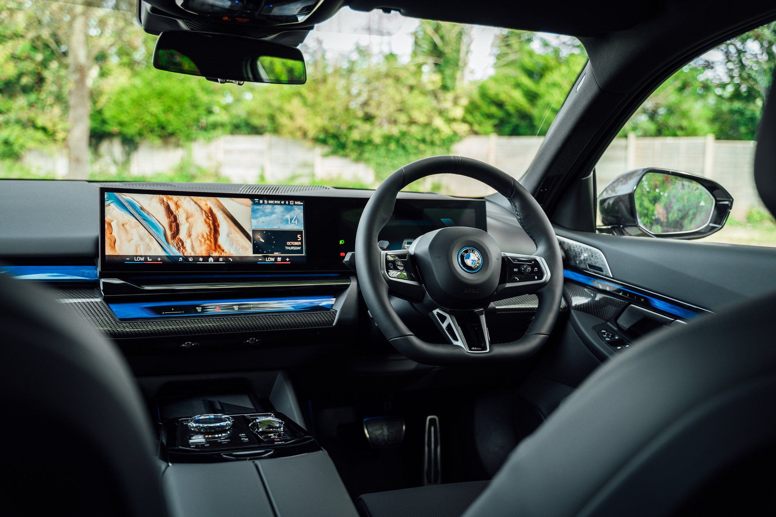 Interior view of a BMW vehicle showing the dashboard, steering wheel, digital display screens, and rearview and side mirrors. The background outside the vehicle has green trees and a wooden fence.