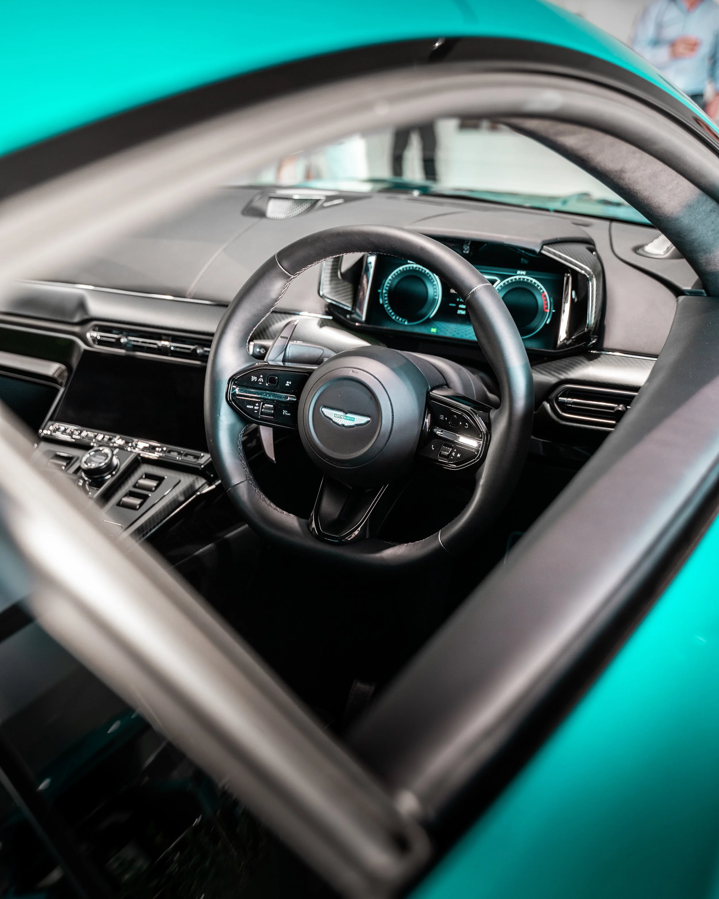 Interior of a car showing the steering wheel, dashboard, and digital instrument cluster. In the background, people are standing and talking.