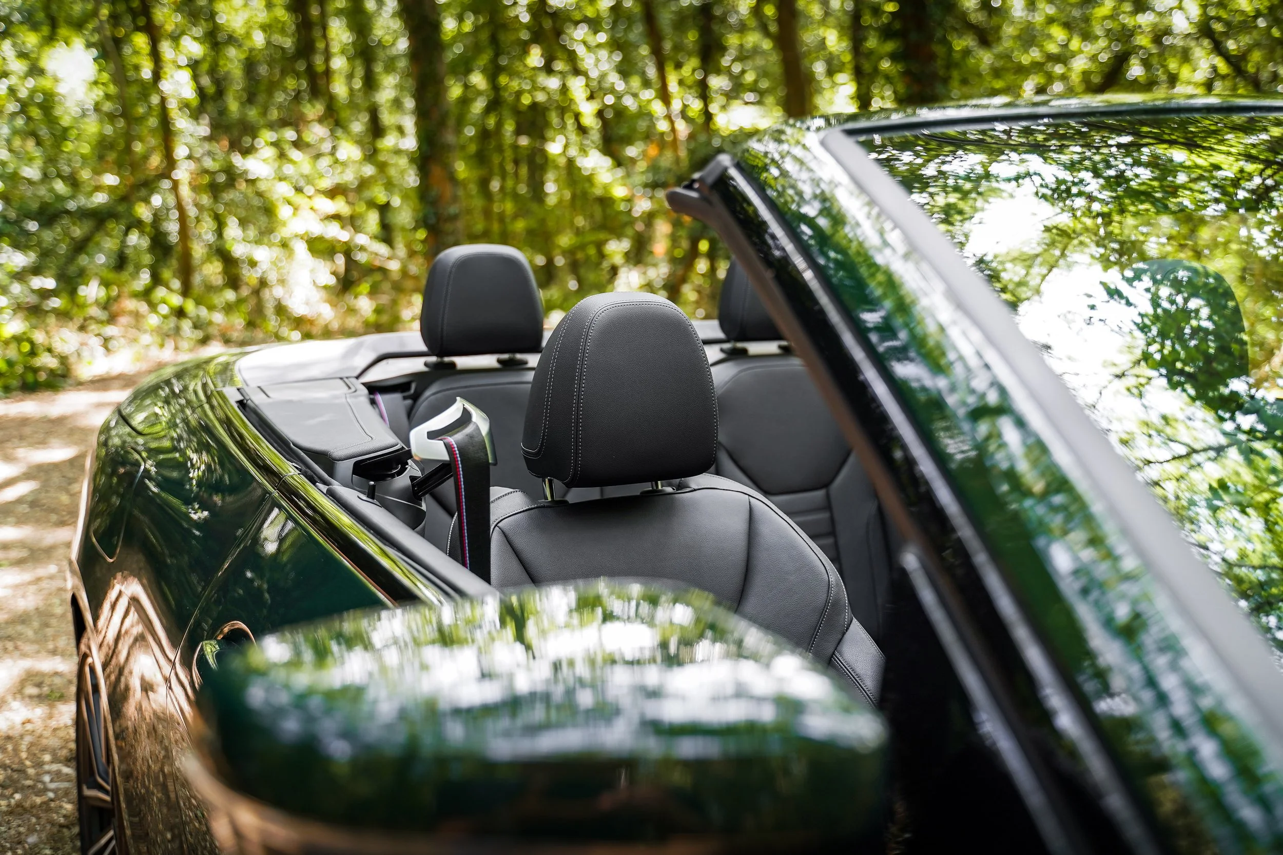 The photo shows the interior of a black convertible car with the top down, parked amidst a forest with dense green trees. The back of the front seats and the windshield reflecting the foliage are visible.