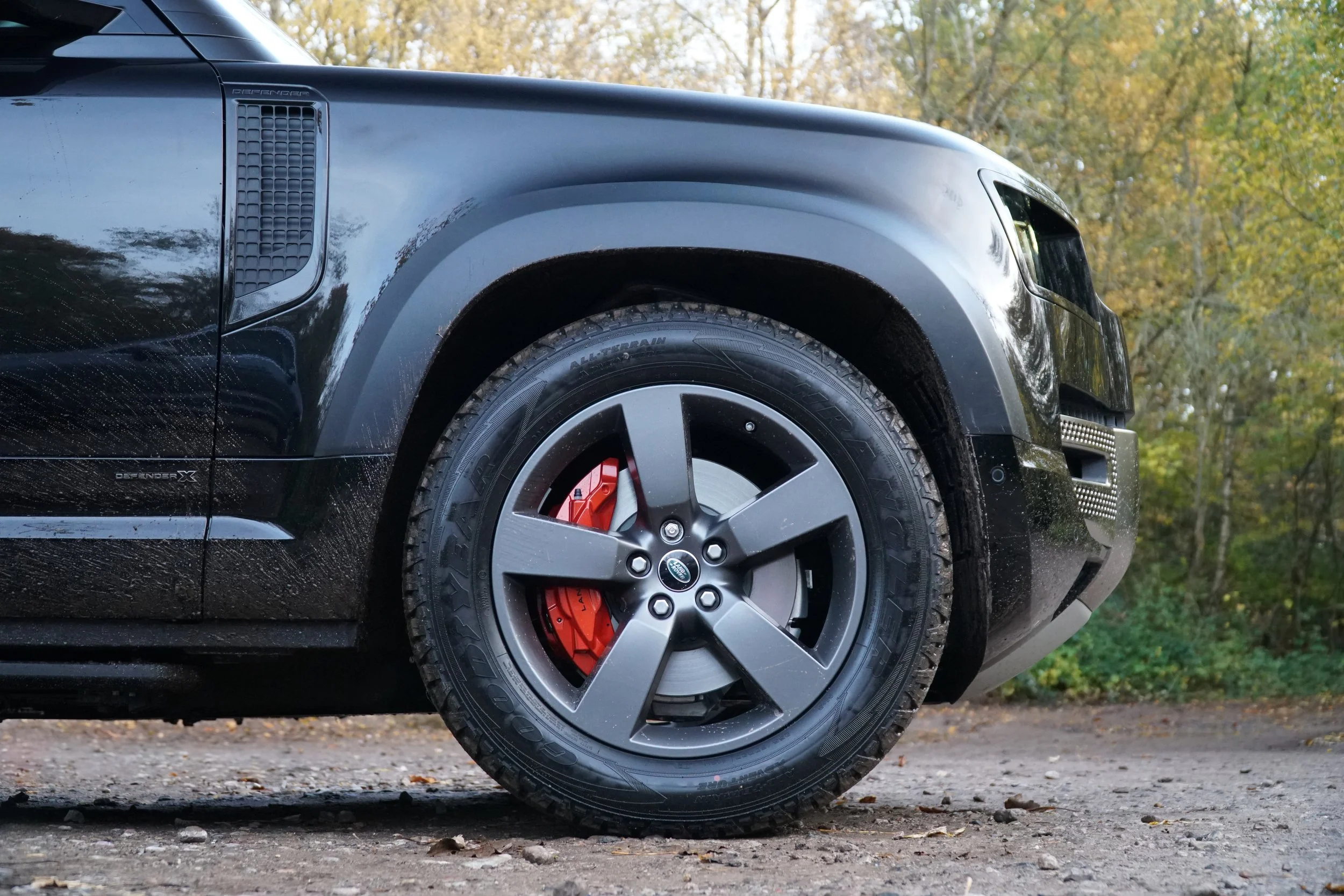 Close-up of a black modern SUV, showing the front right wheel with a black rim and red brake caliper, parked on a gravel ground with autumn trees in the background.