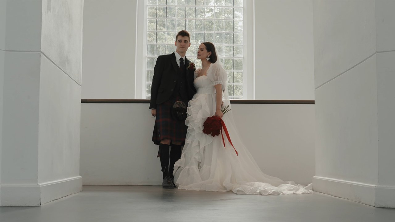 A bride and groom stand together indoors, with the bride holding a bouquet, in front of a large window in a minimalist setting.