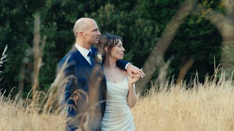 A man in a navy suit and a woman in a white dress stand close together in a field of tall grass with trees in the background.