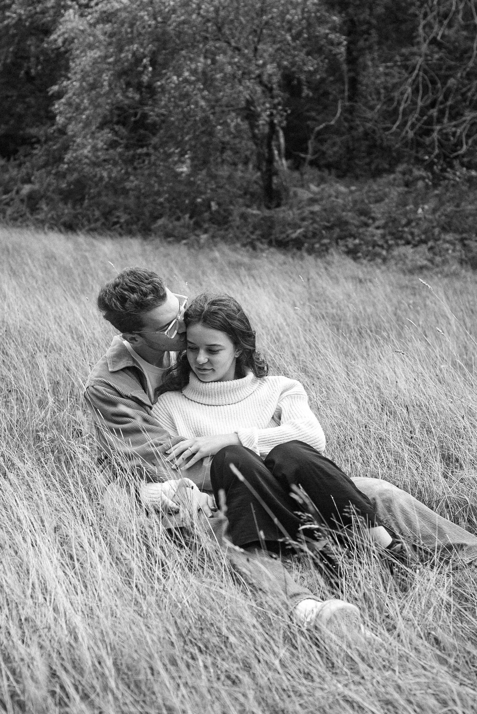 A black and white photo of a couple sitting in a field of tall grass, surrounded by trees in the background. The man is leaning in, kissing the woman on the forehead, who is looking down with a gentle expression.