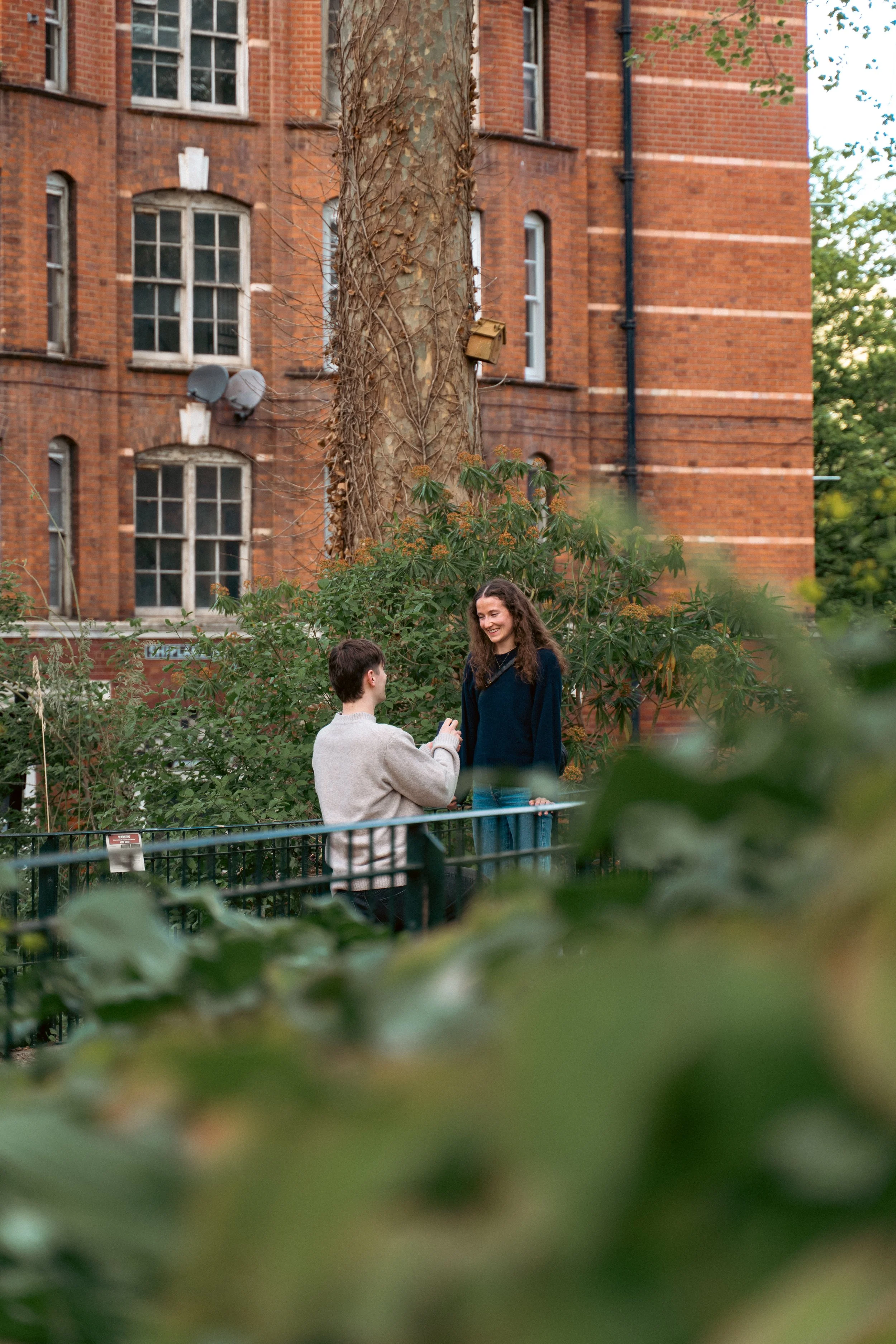 Two young people, a man and a woman, are standing outdoors in a garden area surrounded by green plants. They are smiling at each other, with the man holding a small object, possibly a ring. In the background, there is a large brick building with multiple windows and a tree with some greenery.