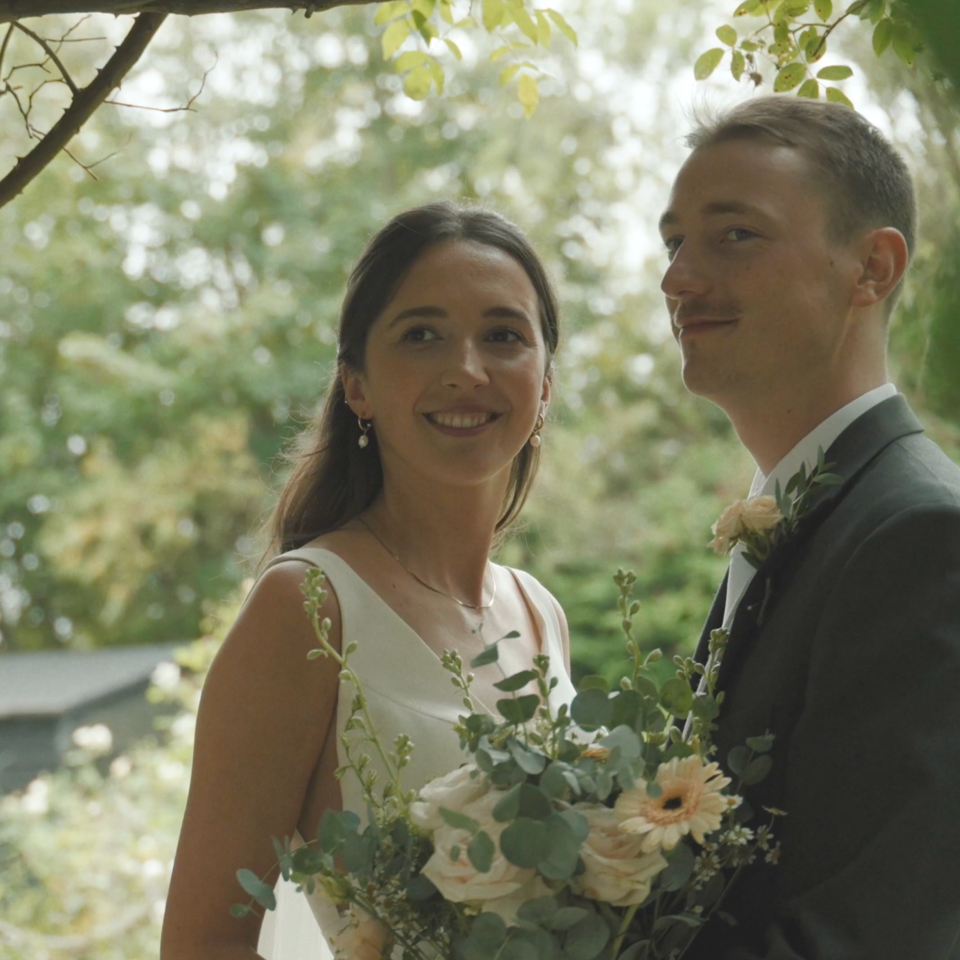 A bride and groom smiling outdoors during a wedding, the bride holds a bouquet of white and peach flowers with greenery, both are dressed in wedding attire, surrounded by trees and natural scenery.