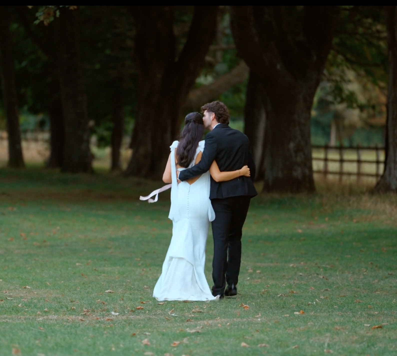 A bride and groom sharing a kiss outdoors in a park with large trees in the background. The bride is wearing a white dress, and the groom is wearing a black suit.